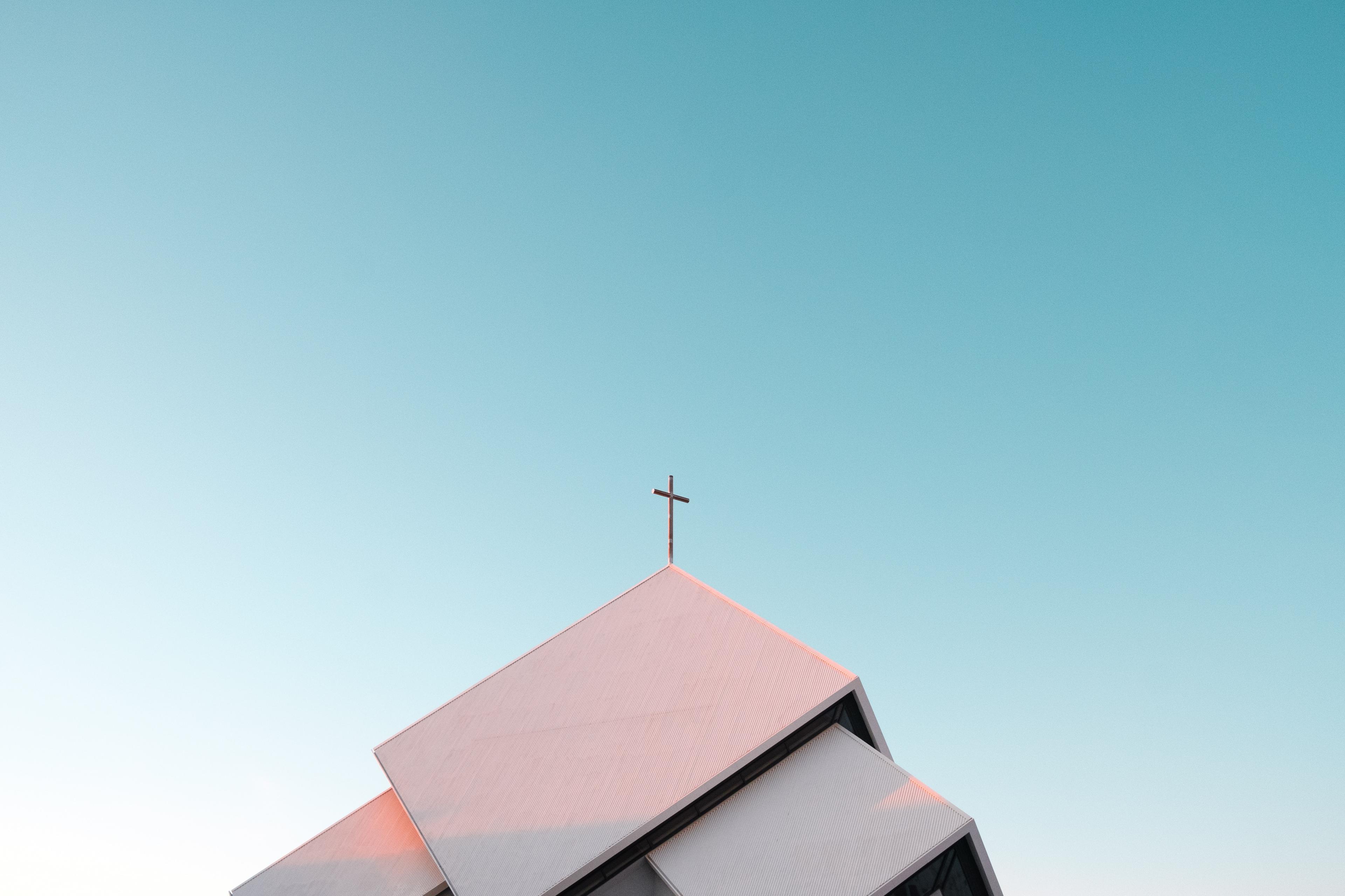 The image features the peak of a modern church with an angular, geometric roofline and a simple cross at the apex, set against a soft gradient sky transitioning from warm pink to pale blue.