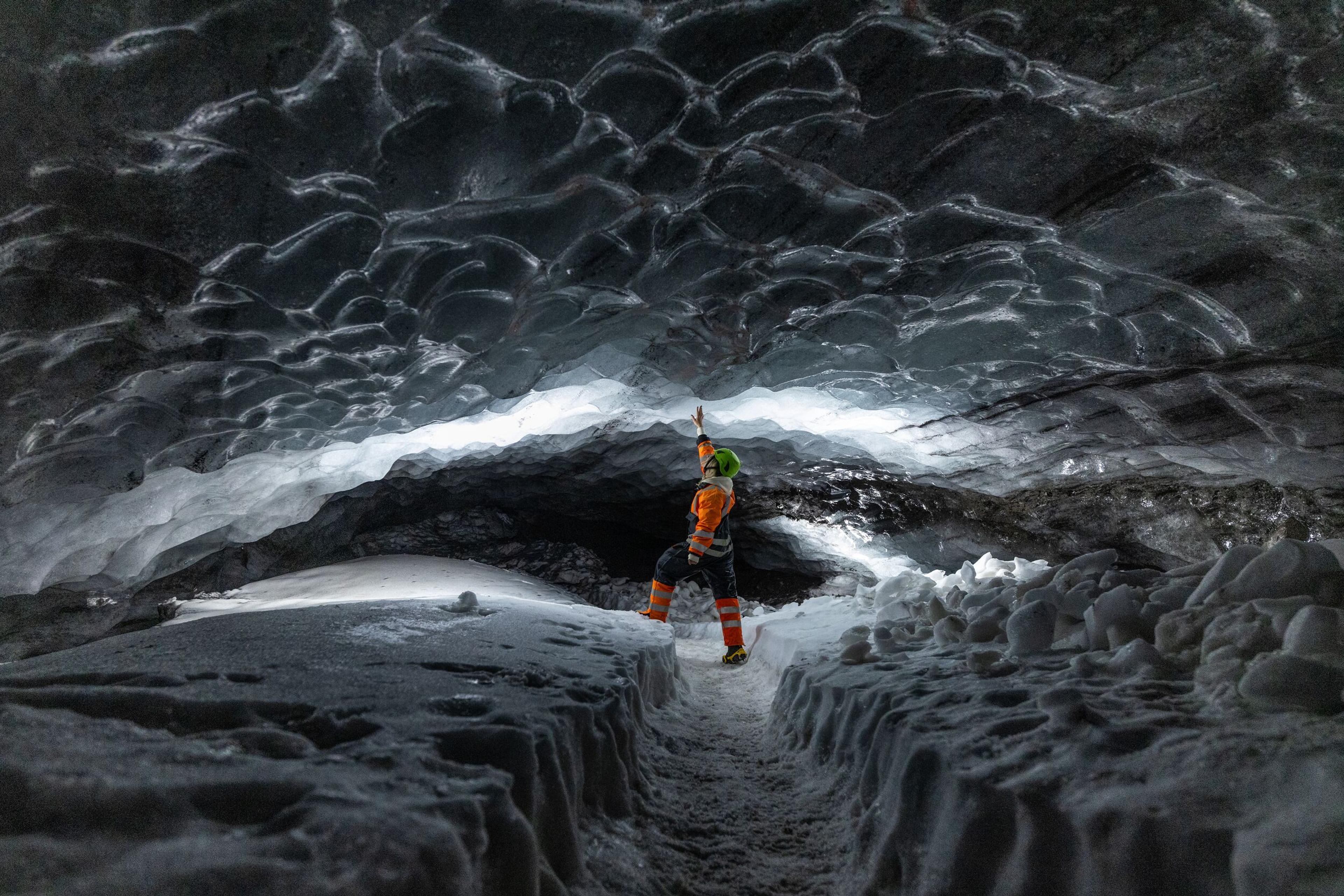 Explorer in orange and black suit pointing at the illuminated ice ceiling inside Askur ice cave, surrounded by snow.