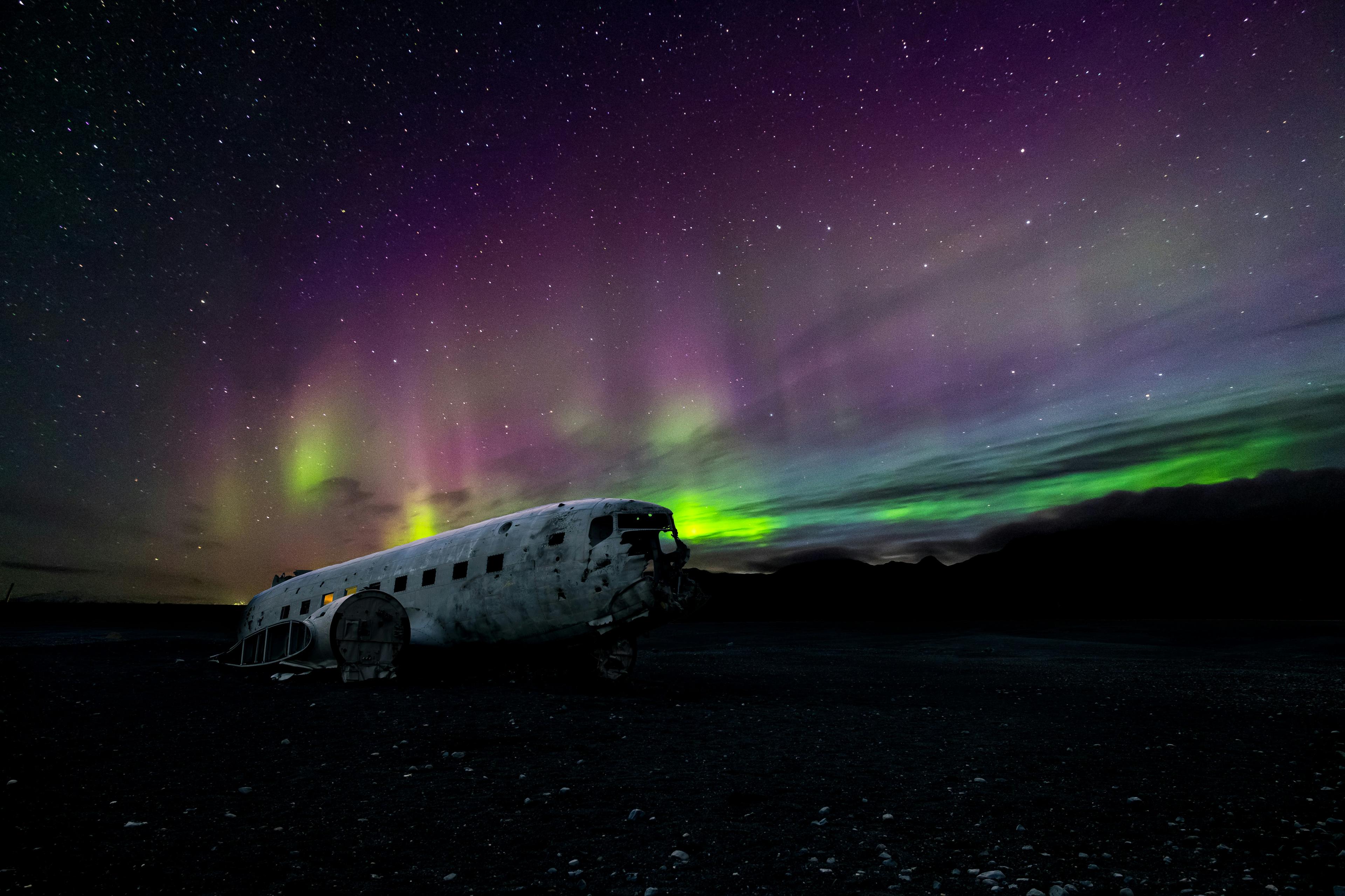 An abandoned airplane sits under a night sky illuminated by vibrant green and purple Northern Lights.