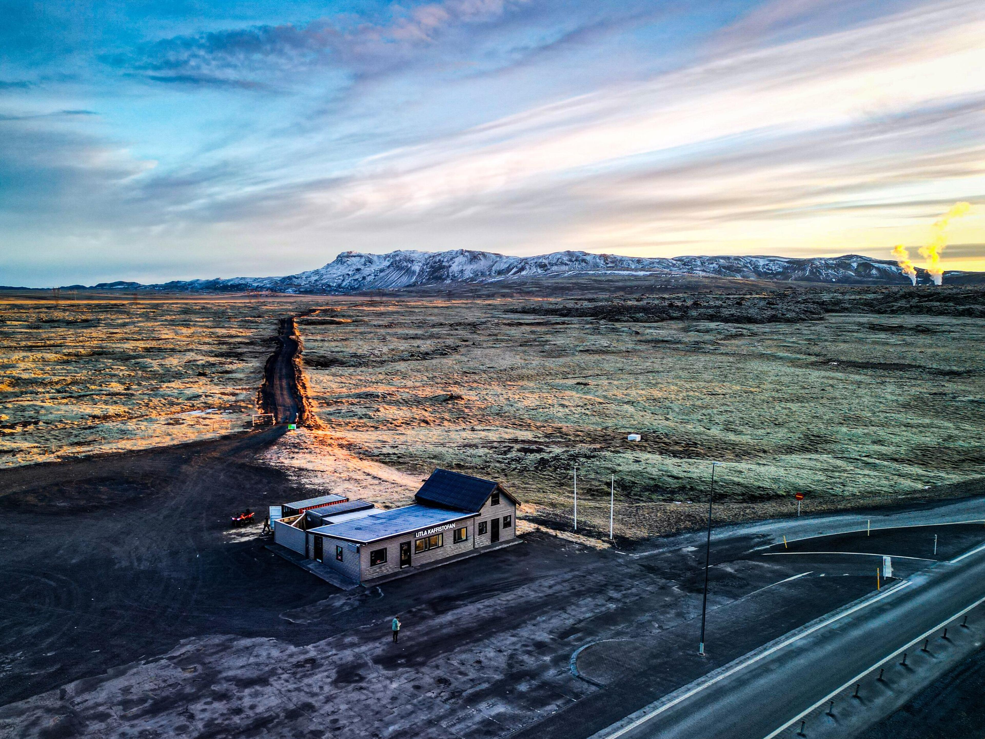 Aerial view of a building with solar panels by a road, bordering a long, dark raised path extending through a vast mossy lava field towards snow-capped mountains and plumes of geothermal steam under a golden-hour sky.