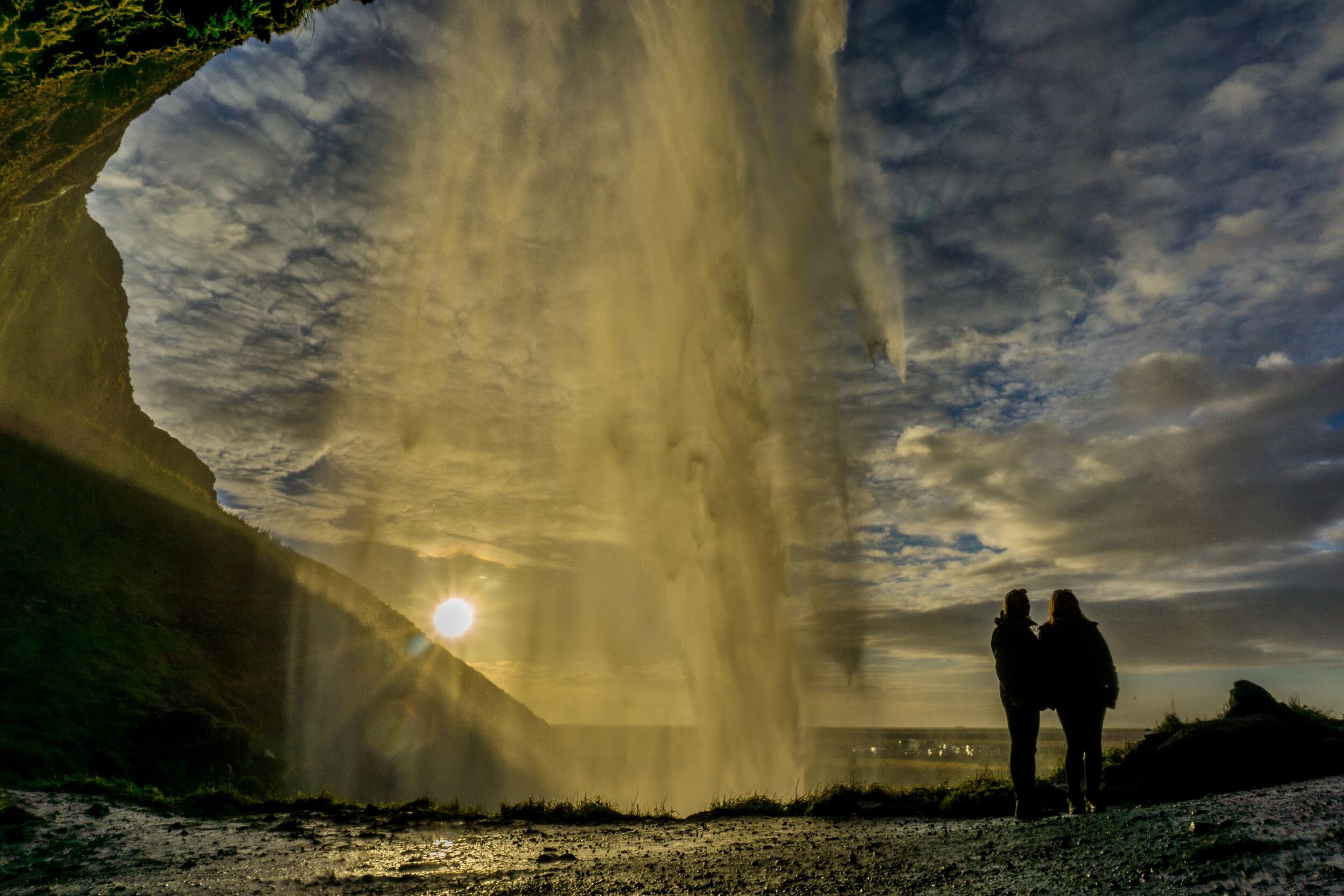 Two people standing behind the cascade with the sun shining trough it, creating a golden hue