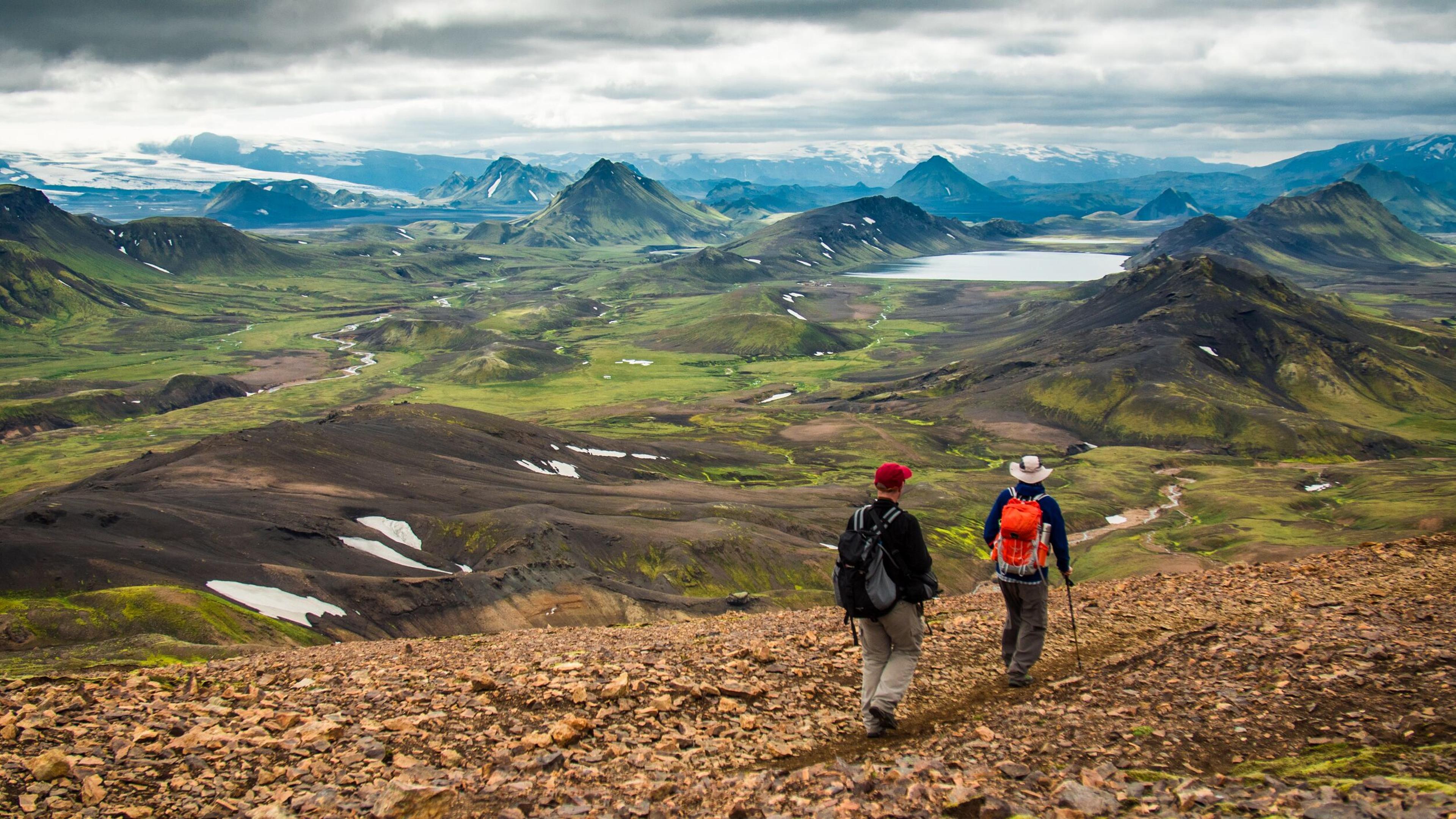 Two hikers on a rocky path overlook a vast landscape of green and dark mountains, a lake, and distant snow.