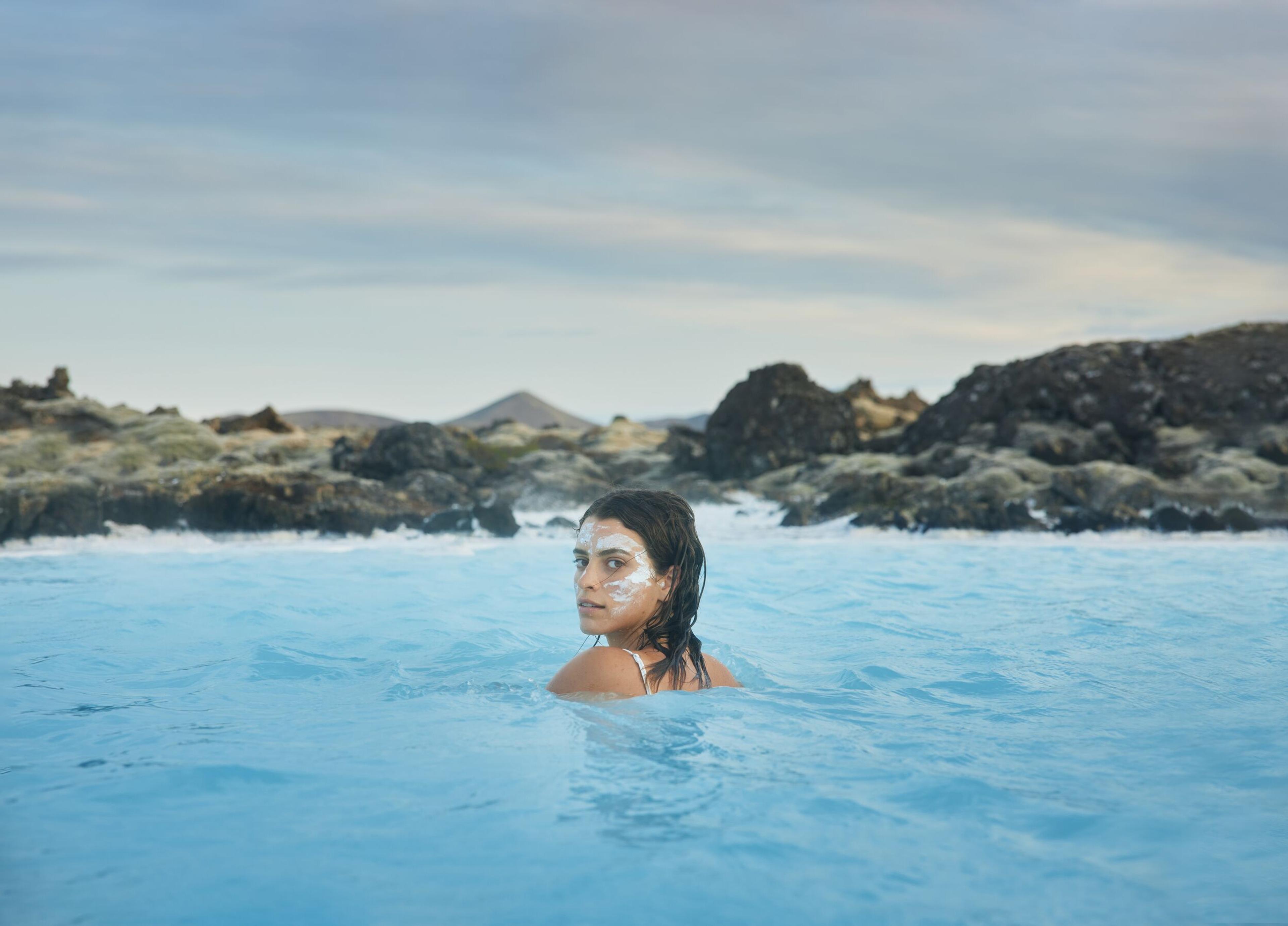 Woman with facial mask enjoys the bright blue waters of the Blue Lagoon in Iceland, surrounded by rocky terrain.