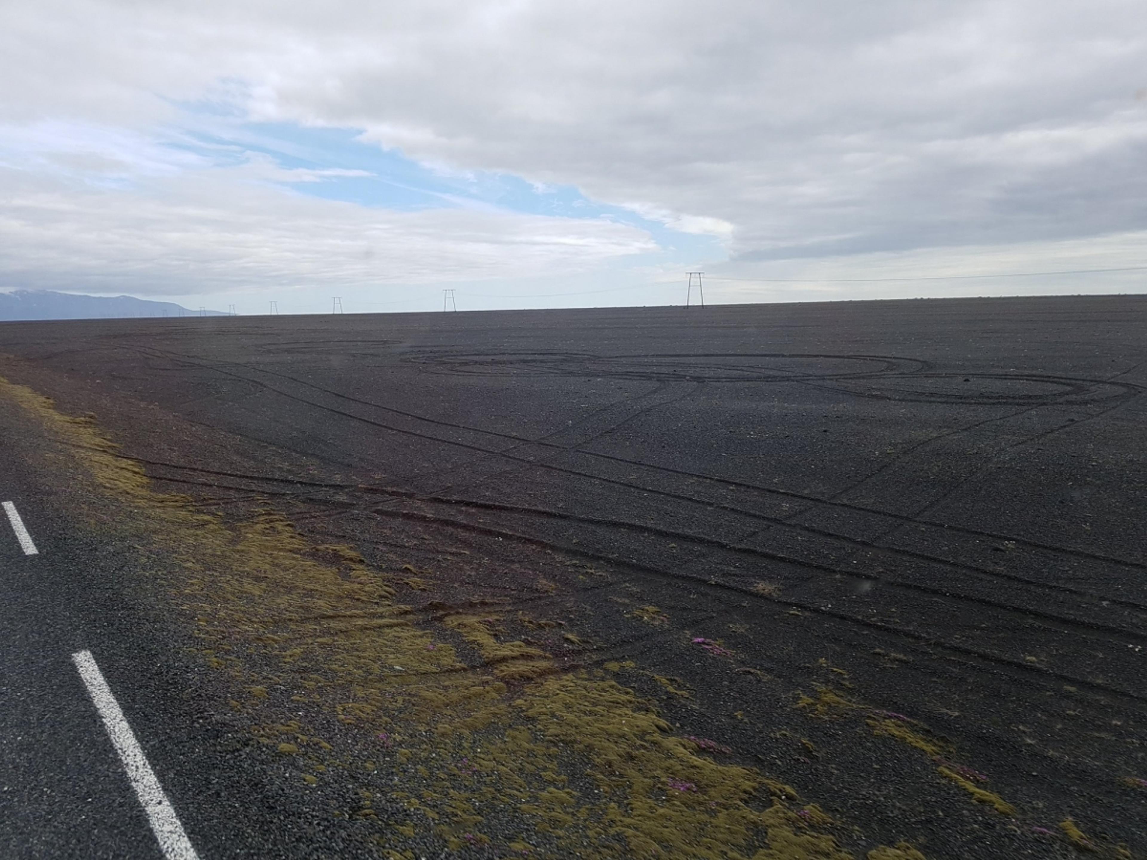 A desolate volcanic landscape stretches beside a road, with dark gravel, tire tracks, and sparse vegetation under a cloudy sky.