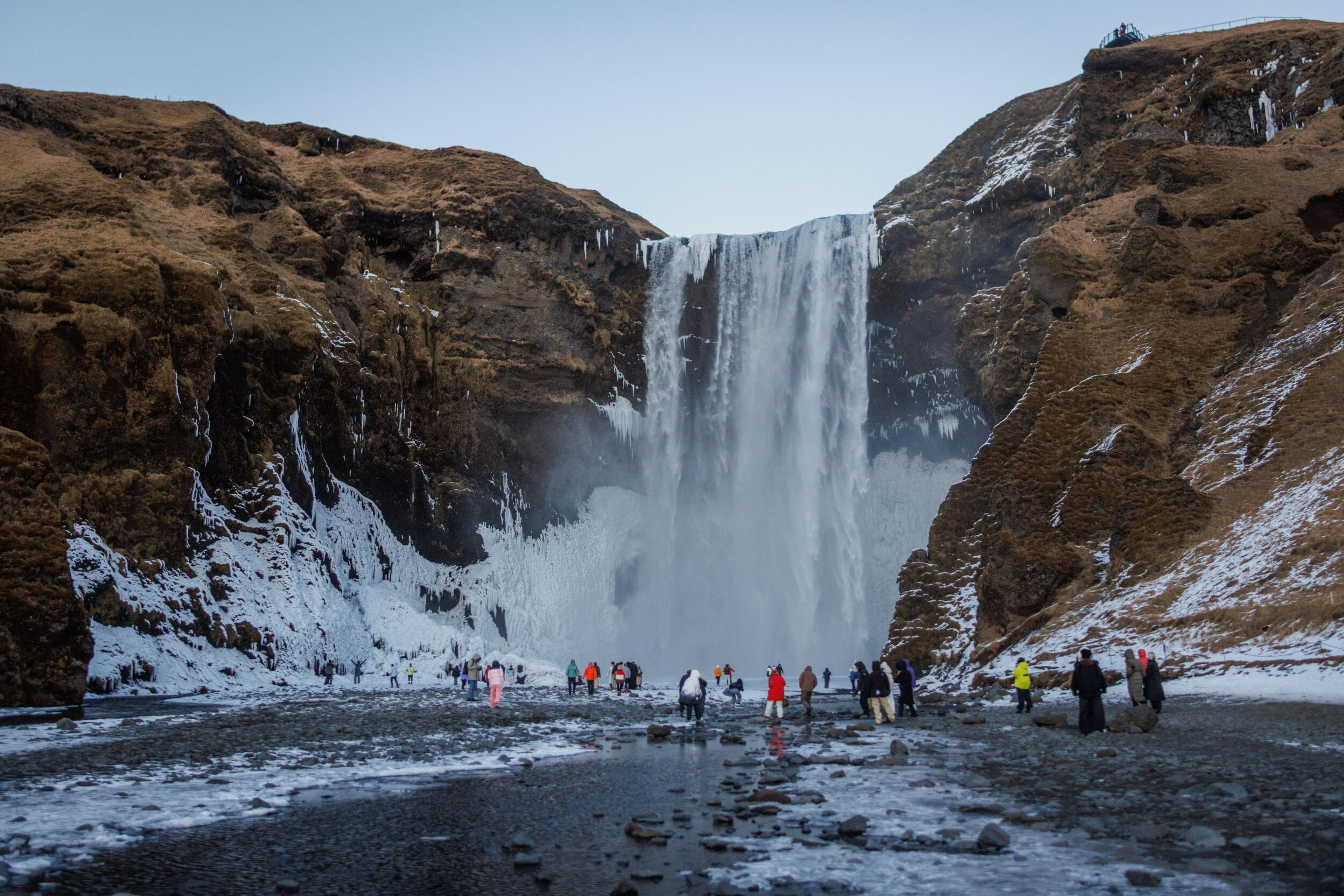 A large waterfall plunges between brown, snow-dusted cliffs, with people standing on the icy ground at its base.