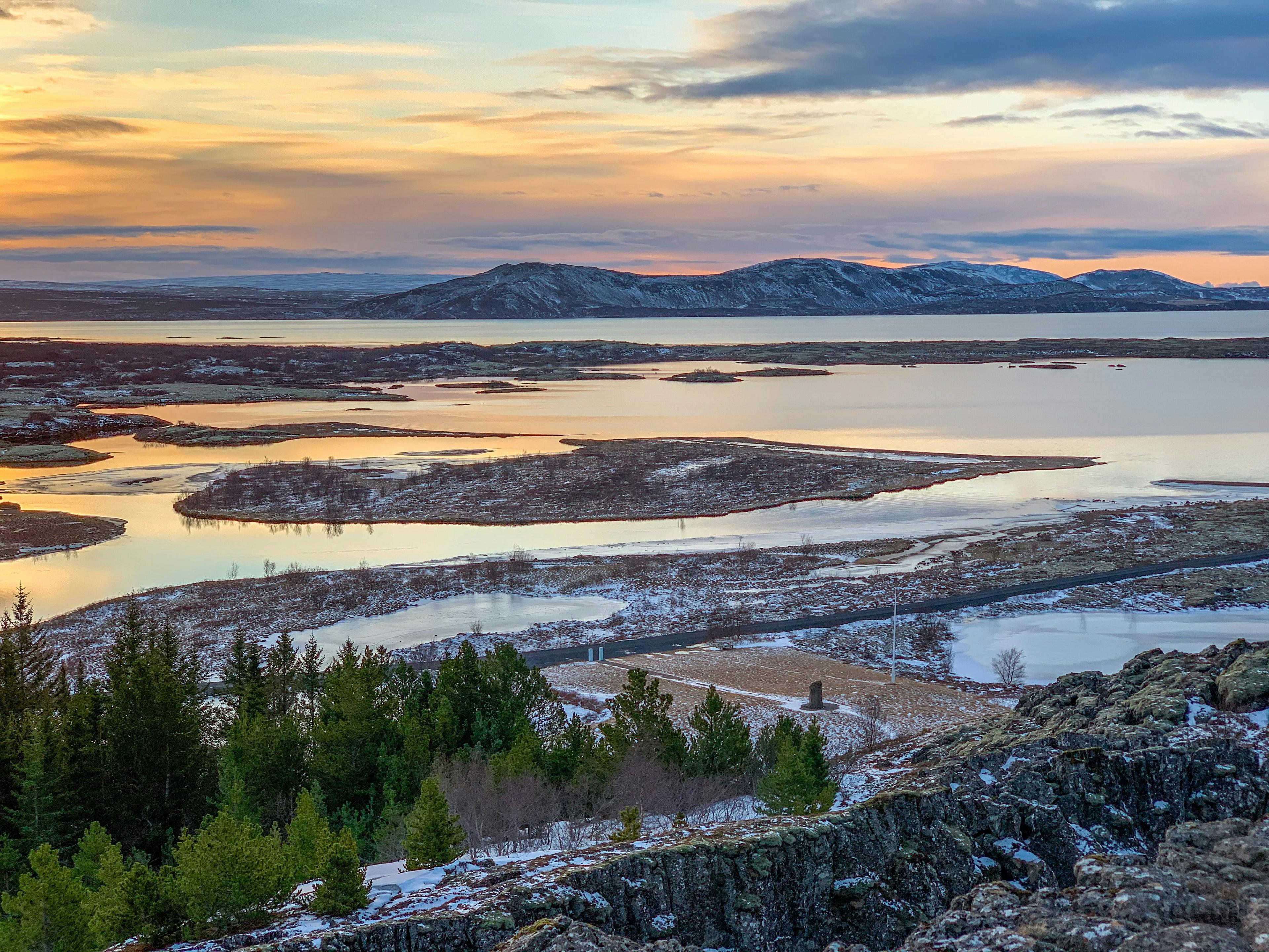 A tranquil sunset view over a vast landscape with reflective waterways, framed by rugged terrain and scattered vegetation, possibly depicting the serene beauty of Þingvellir (Thingvellir) National Park in Iceland.