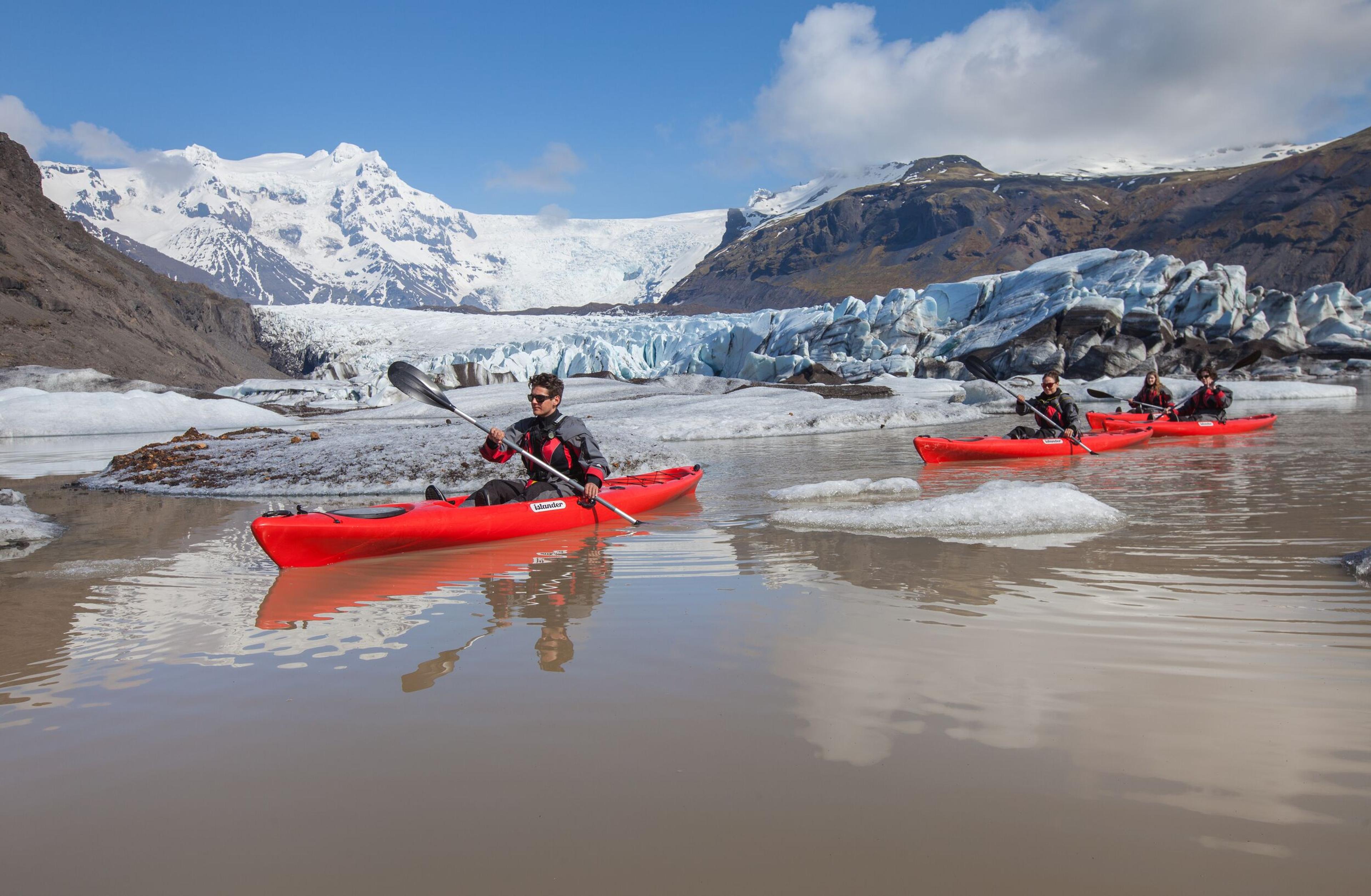 Kayakers in orange kayaks paddling near massive blue glaciers in Iceland.