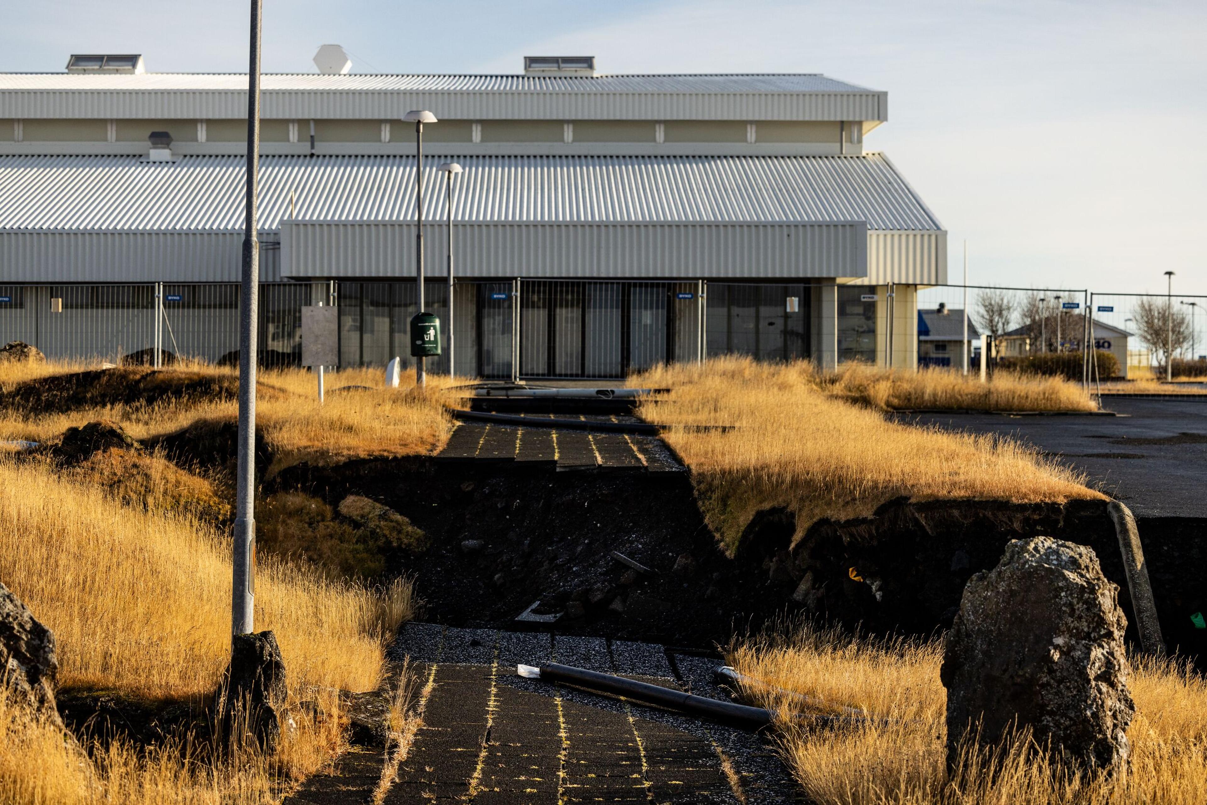 A large industrial building in Grindavík stands behind a cracked pathway, where the ground has split due to recent volcanic activity on the Reykjanes Peninsula.