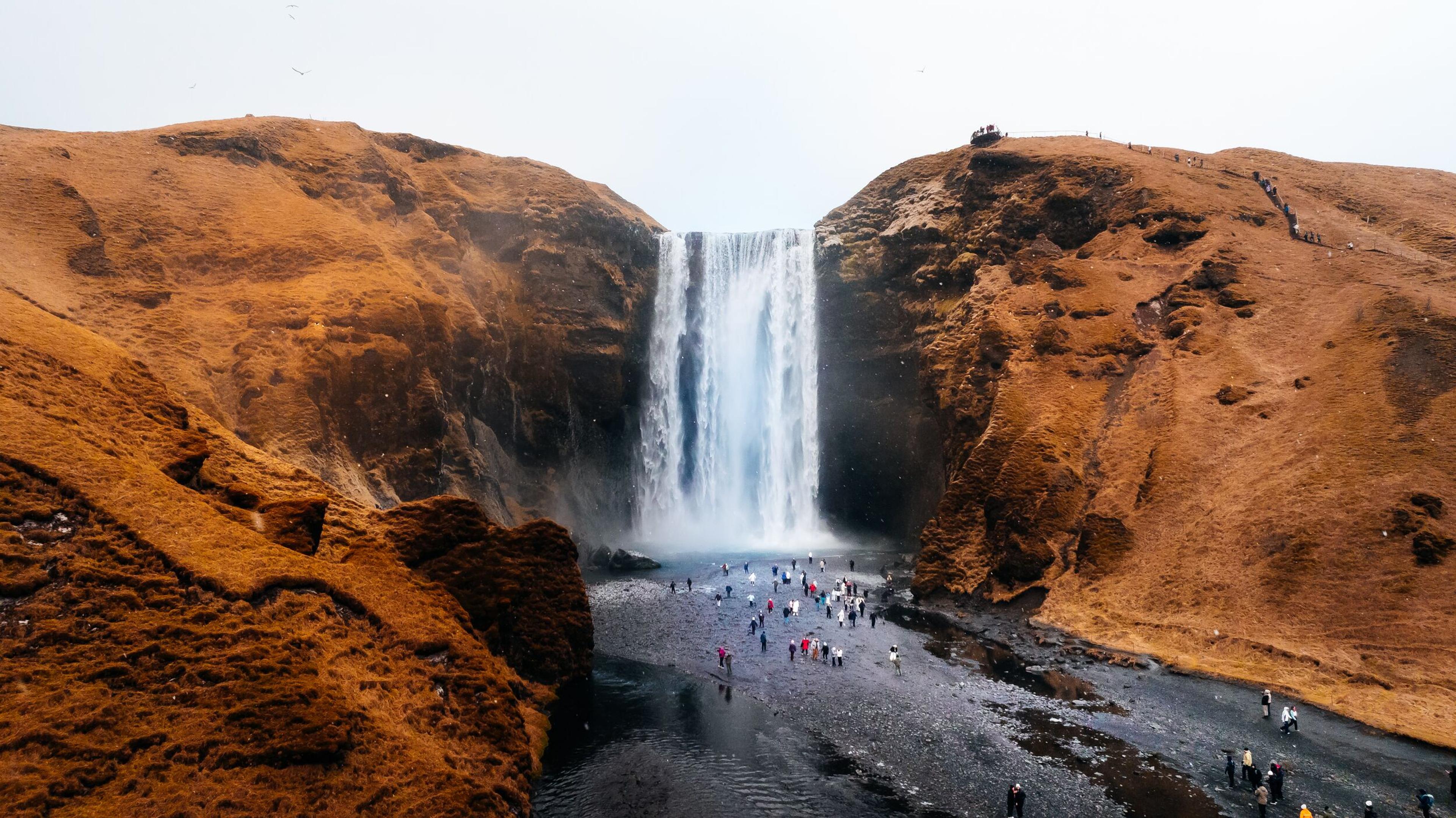 A tall waterfall cascades between two brown, grassy hills, with many people gathered at its base.