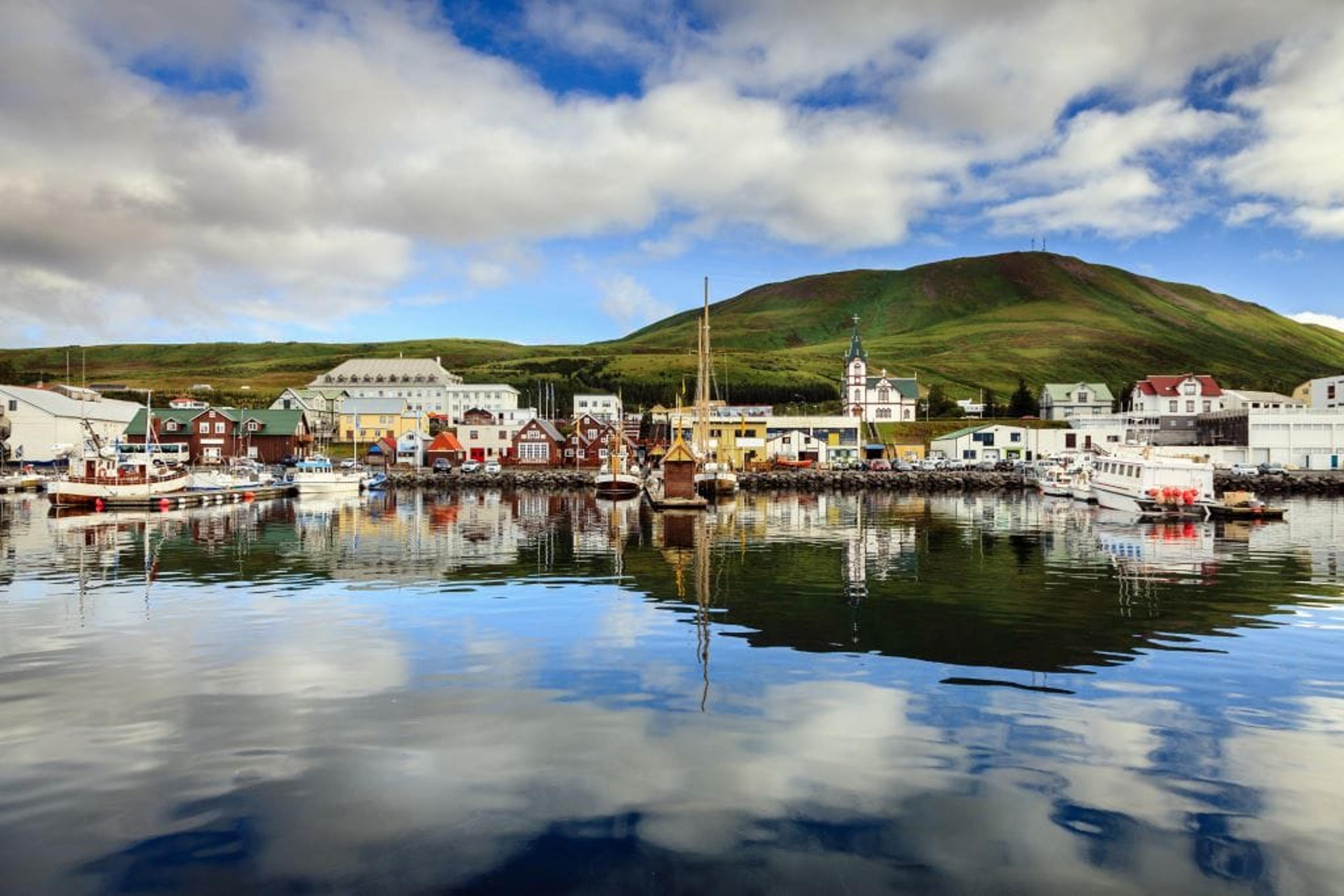 A colorful harbor town with boats reflecting in calm water, backed by a large green hill under a cloudy sky.