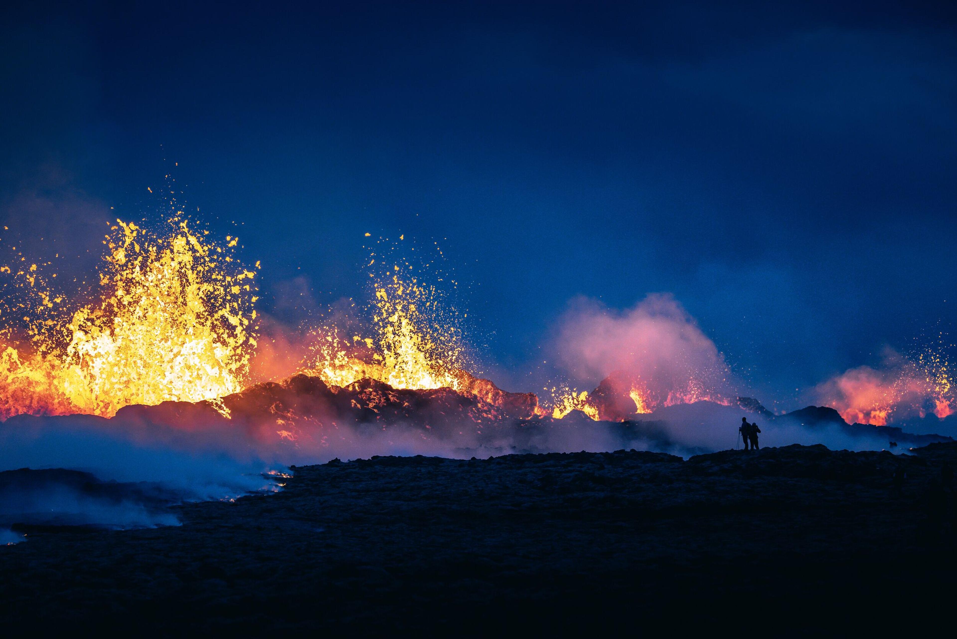 Silhouetted figure against fiery volcanic eruption at twilight, with intense blue smoke and orange lava flames.