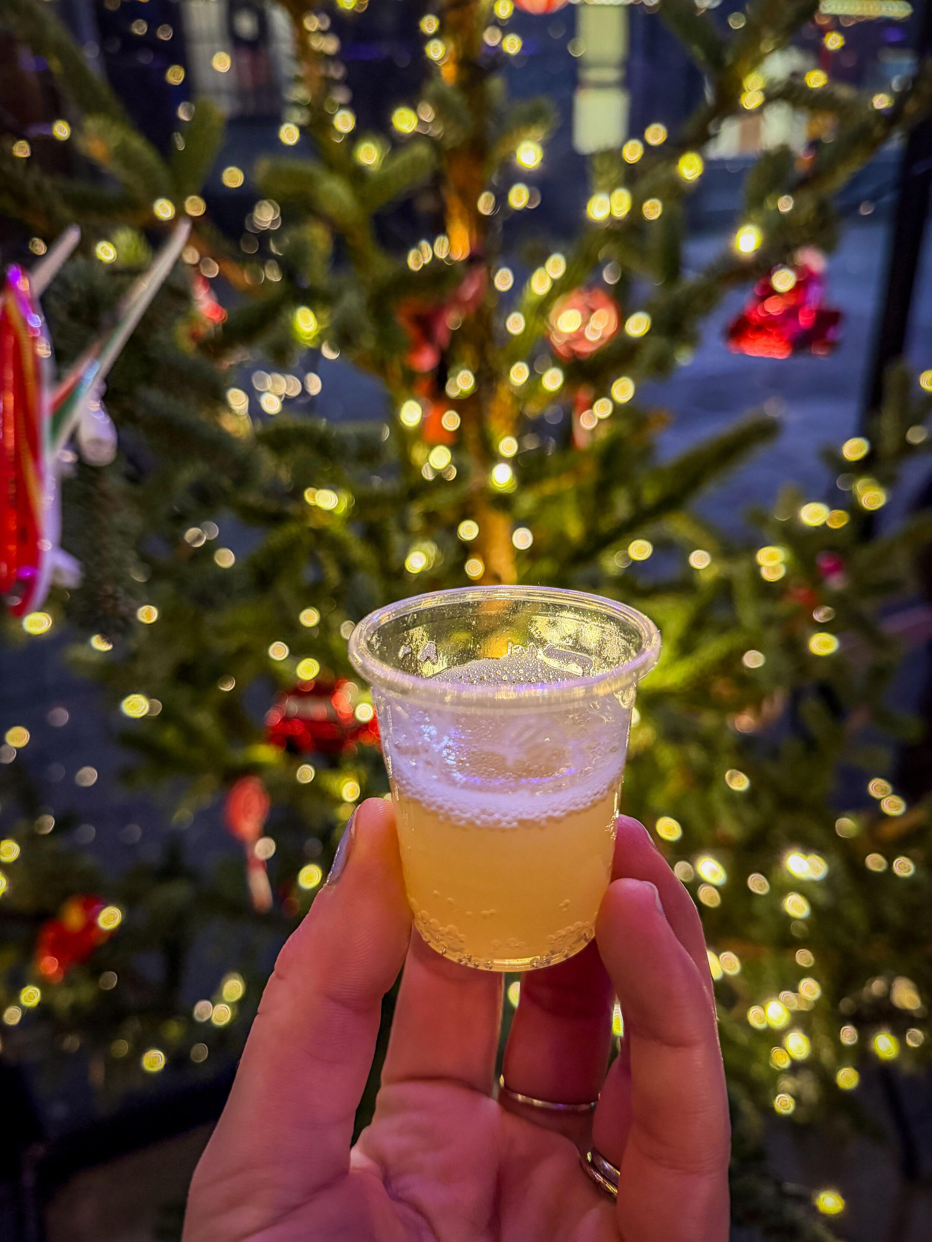 Hand holding a small cup of bubbly yellow drink in front of a blurred, lit Christmas tree.