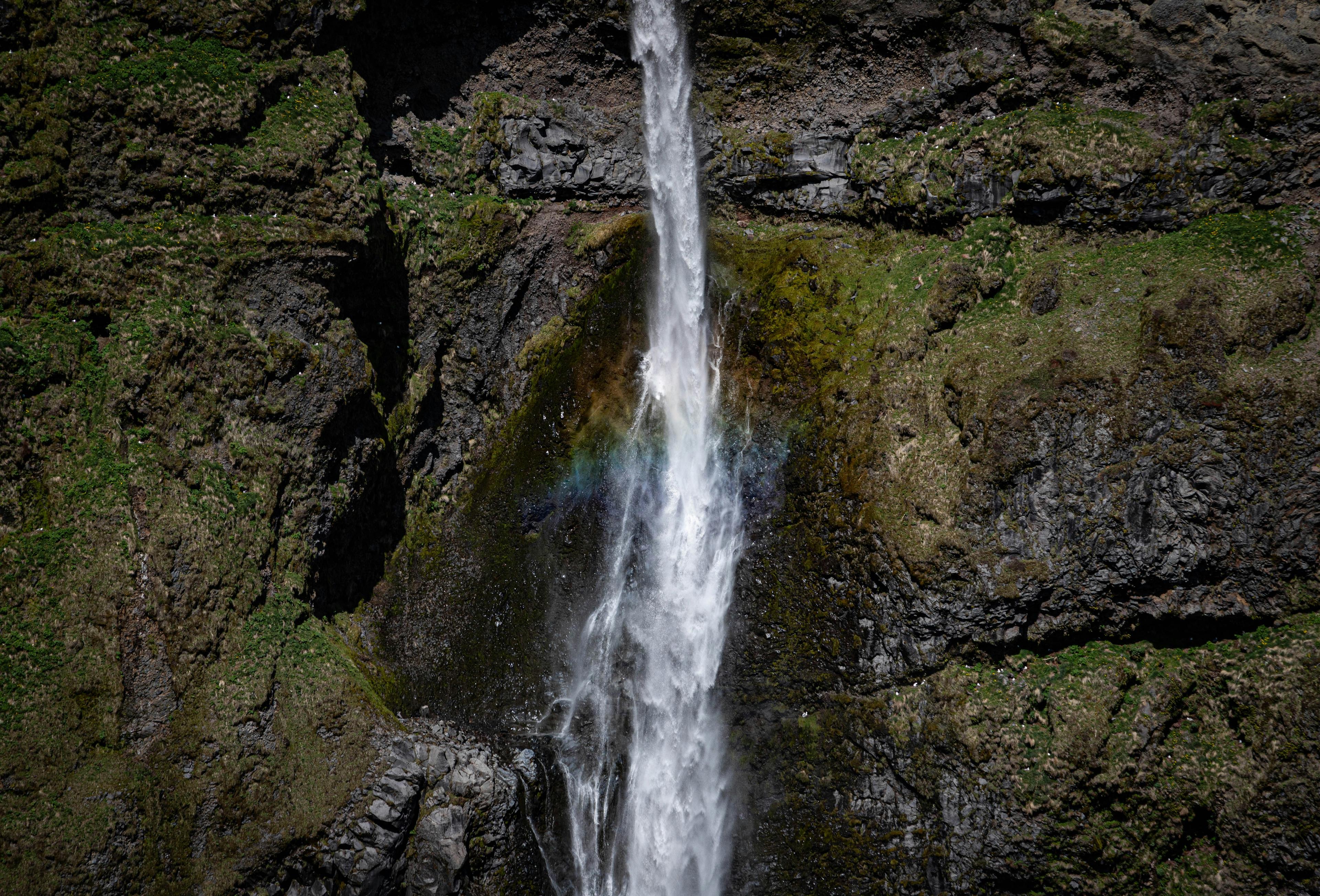 Close-up of a waterfall in Mulagljufur Canyon, capturing the water's descent and a rainbow forming in the spray against dark rocky cliffs.