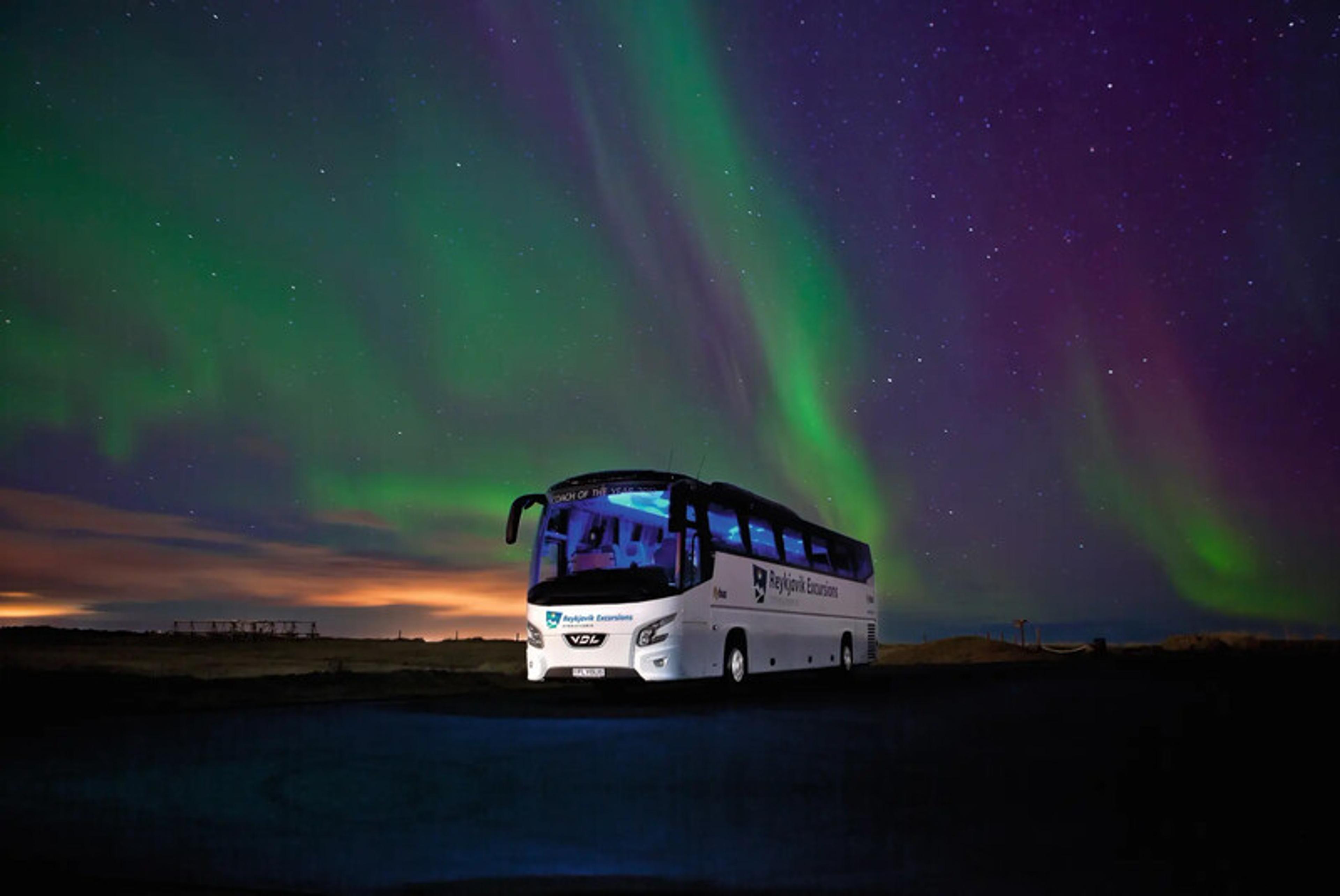 A tour bus is parked under a night sky illuminated by the vivid colors of the Northern Lights, with shades of green and purple dancing across the horizon.