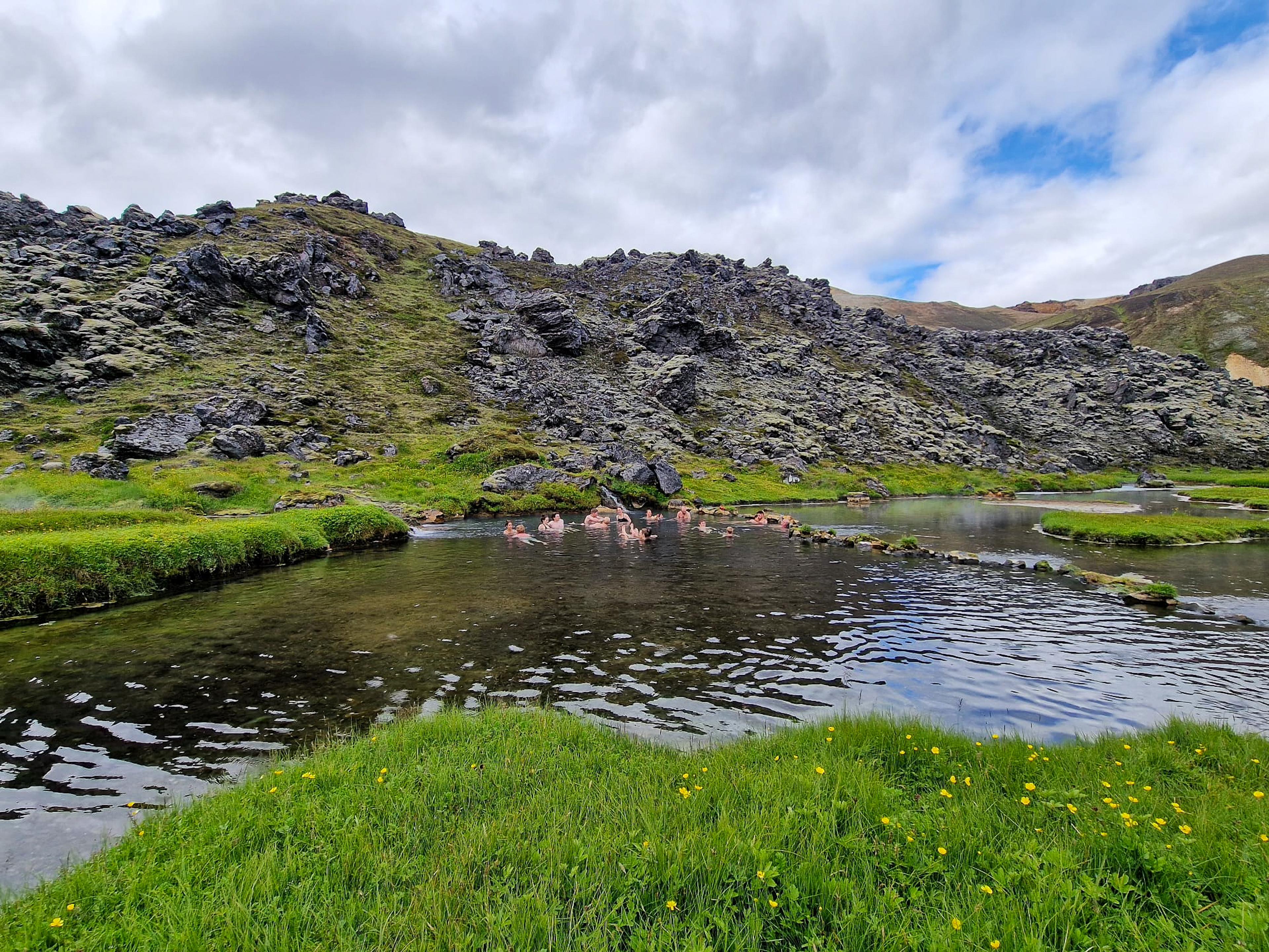 People bathing in a clear stream surrounded by vibrant green grass and dark, moss-covered rocky hills.