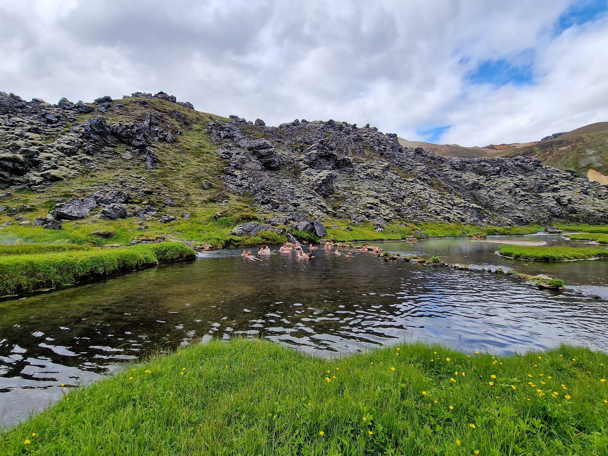 People bathing in a clear stream surrounded by vibrant green grass and dark, moss-covered rocky hills.