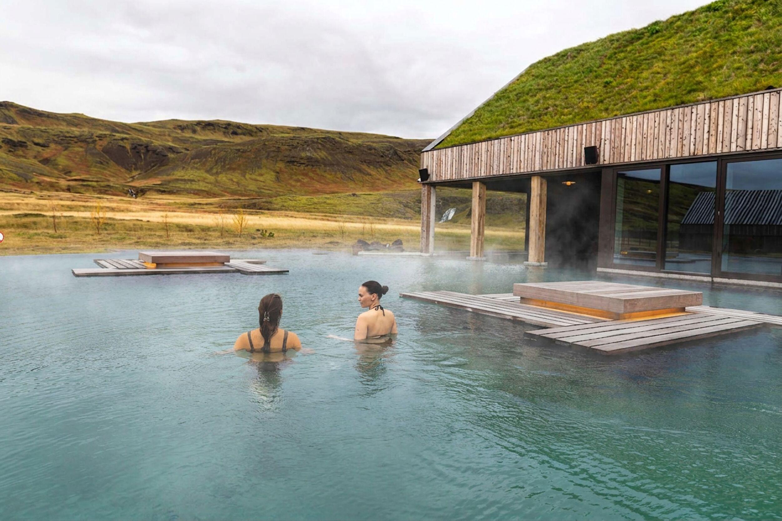 Two women in a steaming outdoor geothermal pool, with a modern building featuring a grass roof and rolling hills in the background.