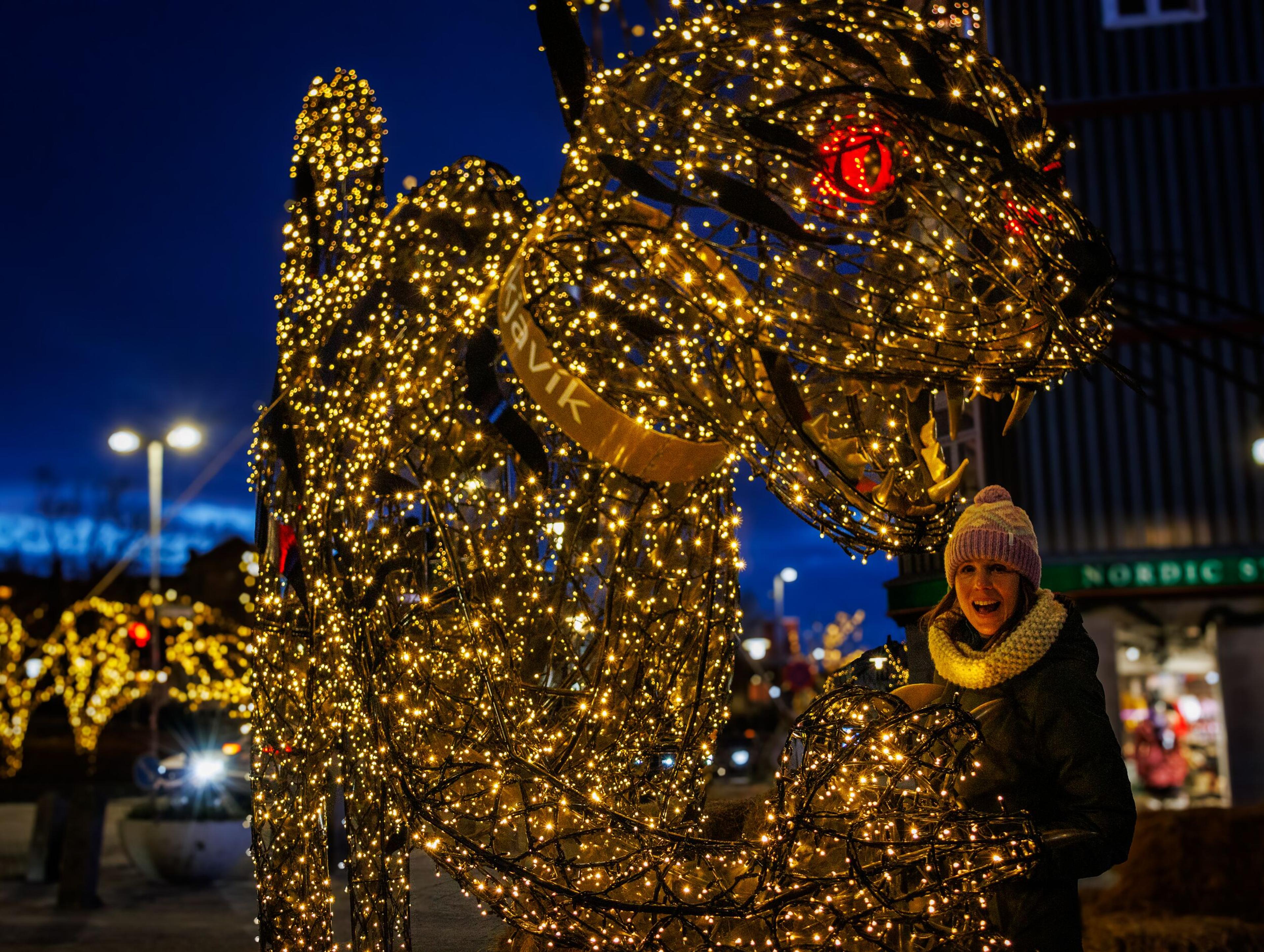 A smiling woman stands beside a large dragon sculpture with golden lights and red eyes, at night in Reykjavik.