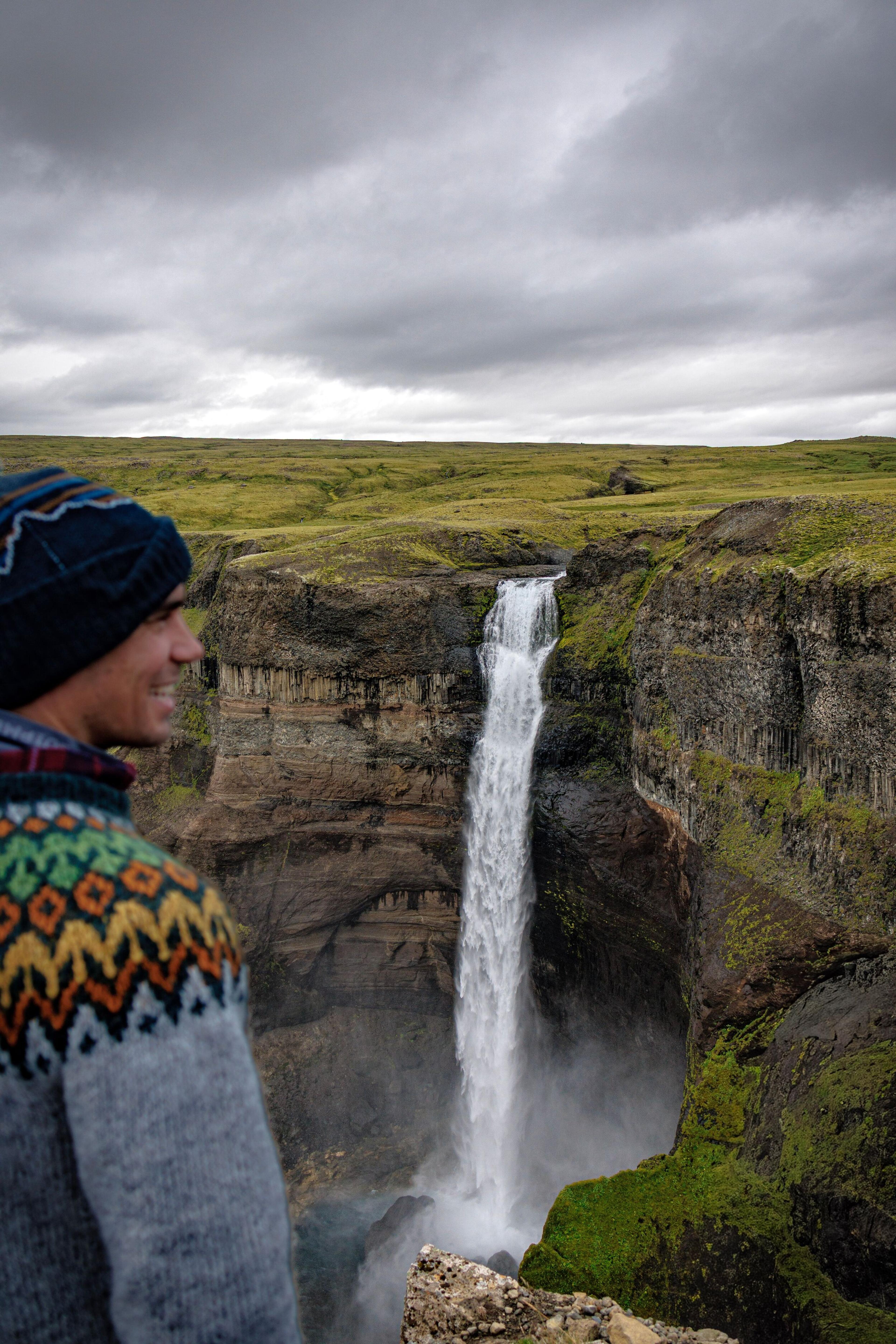 A smiling man in a patterned sweater and beanie looks at a tall waterfall cascading into a rocky canyon.