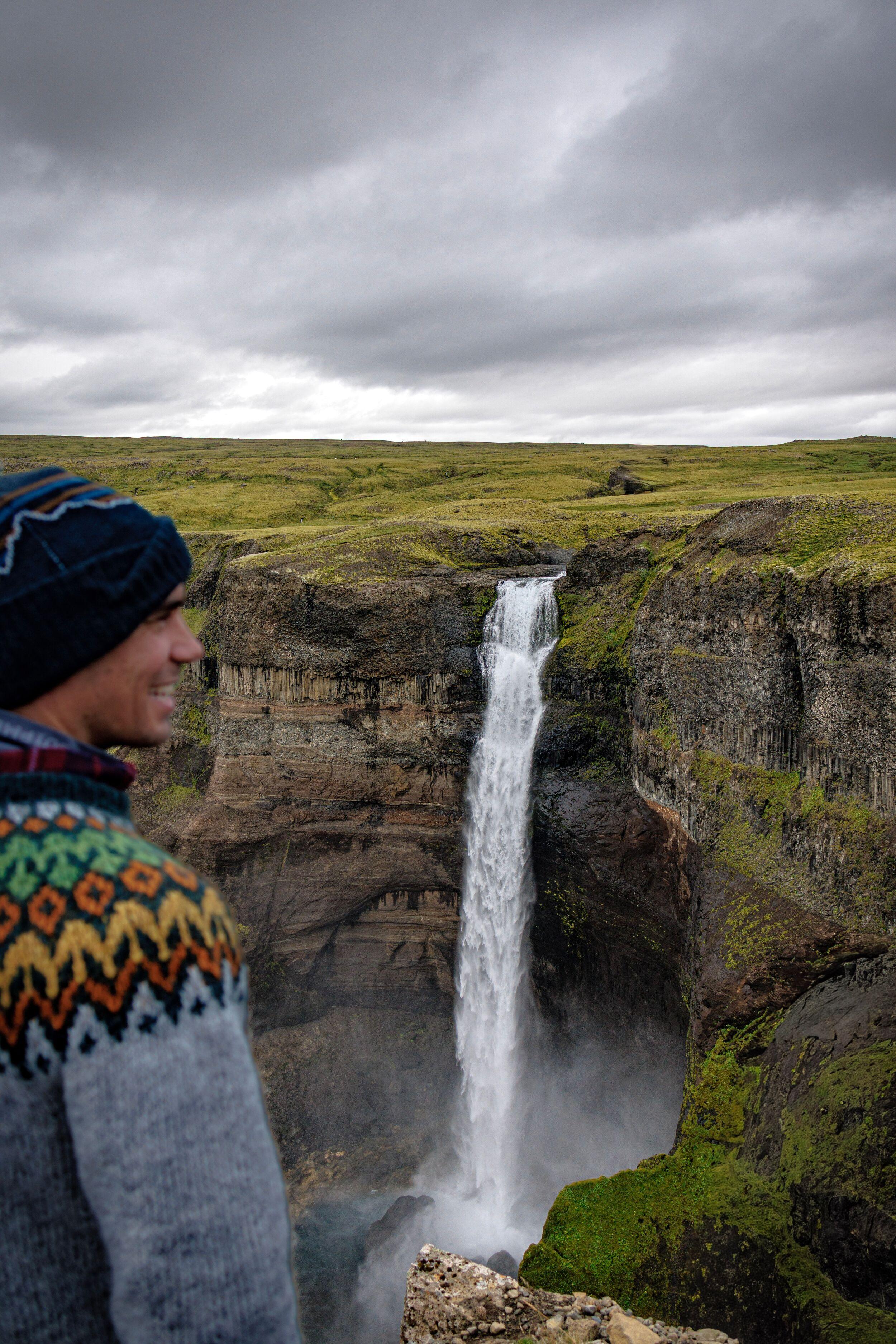 A smiling man in a patterned sweater and beanie looks at a tall waterfall cascading into a rocky canyon.