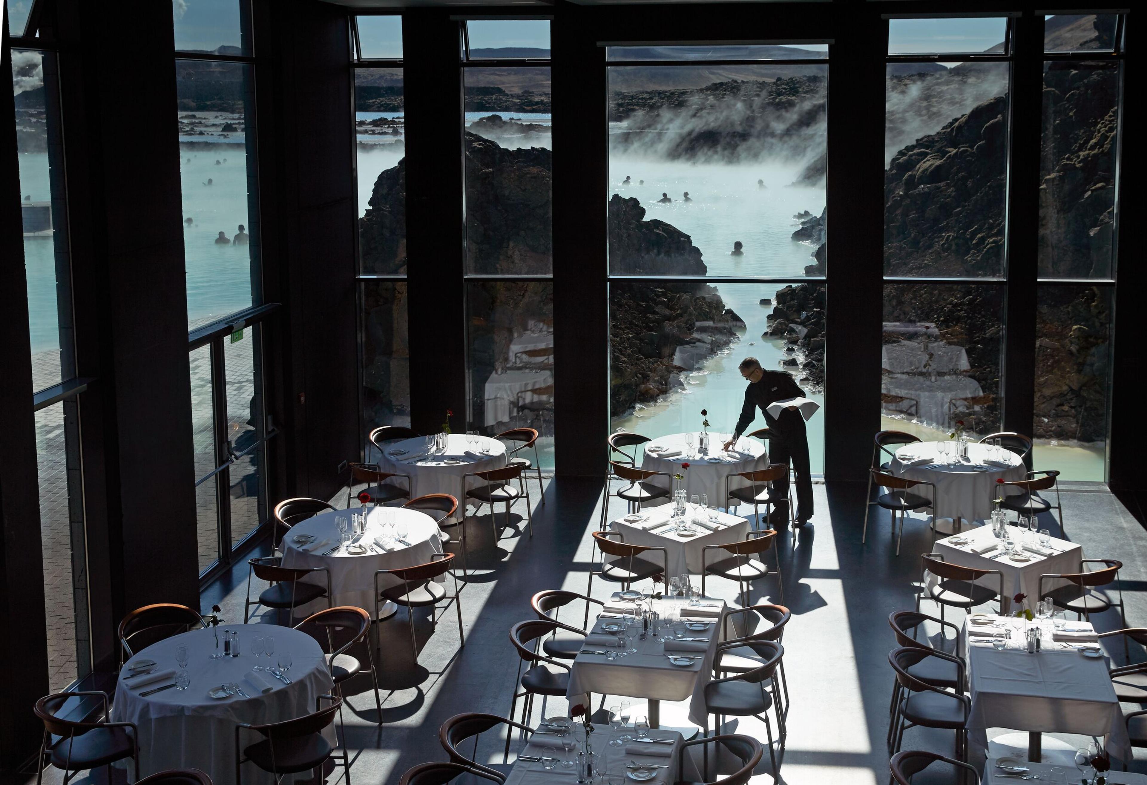 A waiter sets up tables for dinner service, while behind large windows, guests enjoy a swim in the lagoon.