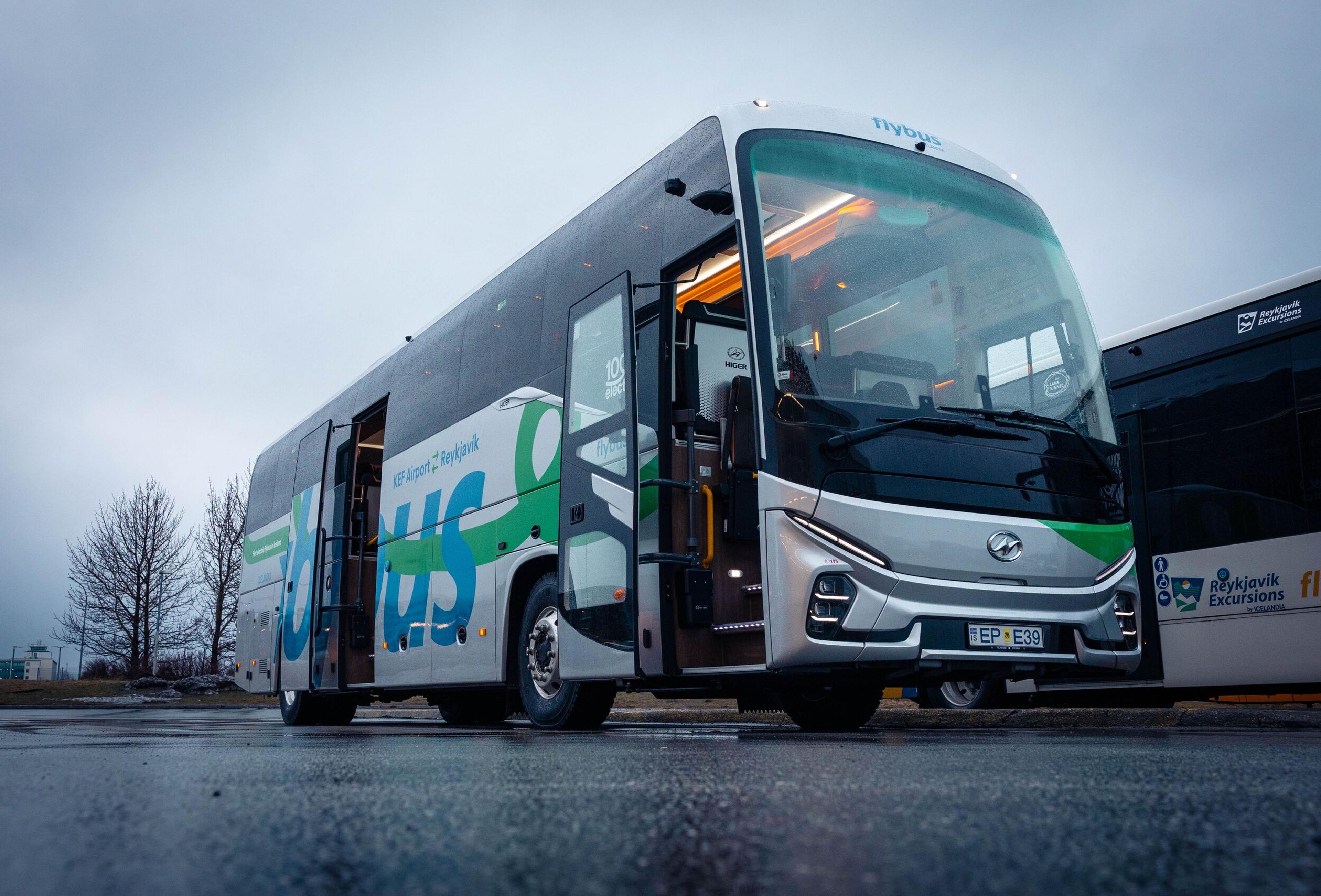 A modern silver and green Flybus with its doors open is parked on wet asphalt under an overcast sky.