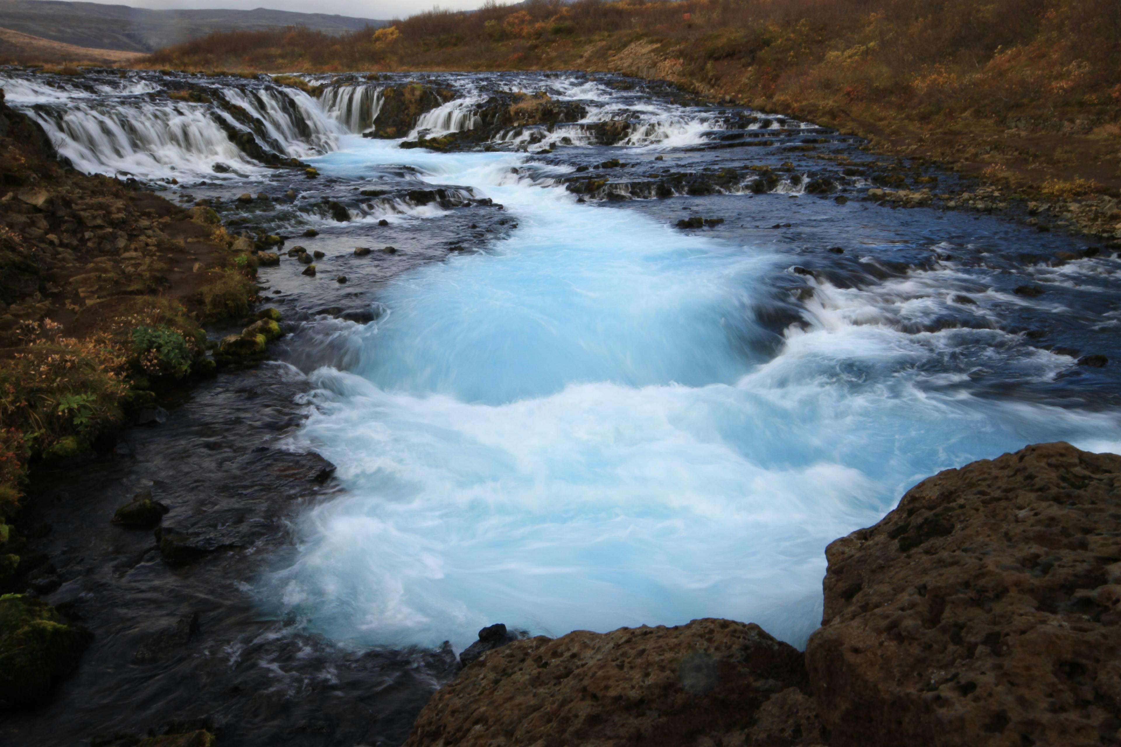 A vivid blue stream rushing through a multi-tiered waterfall, framed by rugged terrain and autumn-hued vegetation, capturing the raw beauty of Iceland's dynamic waterways.