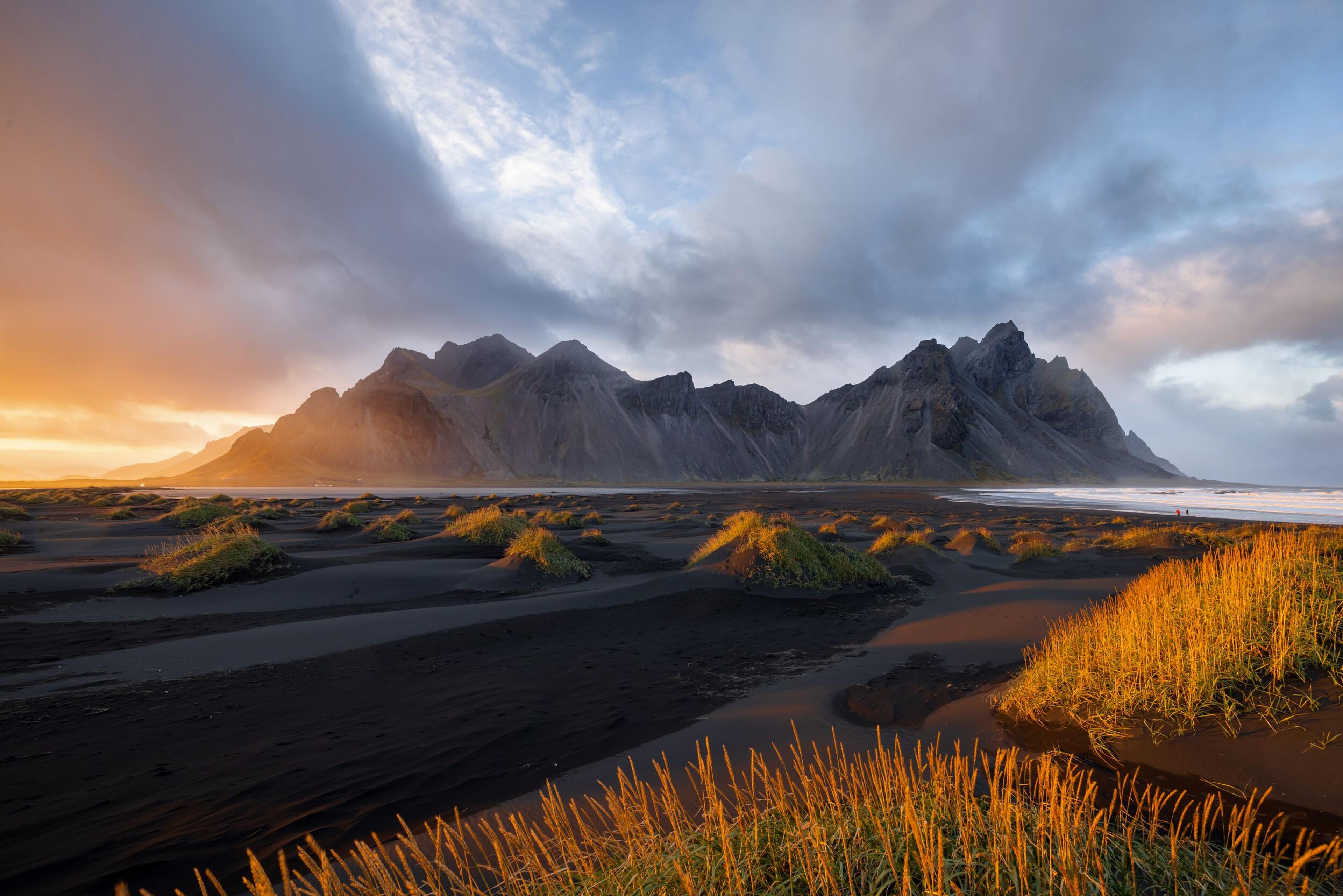 Black sand beach and golden grass foreground with jagged dark mountains under a cloudy sunset sky.