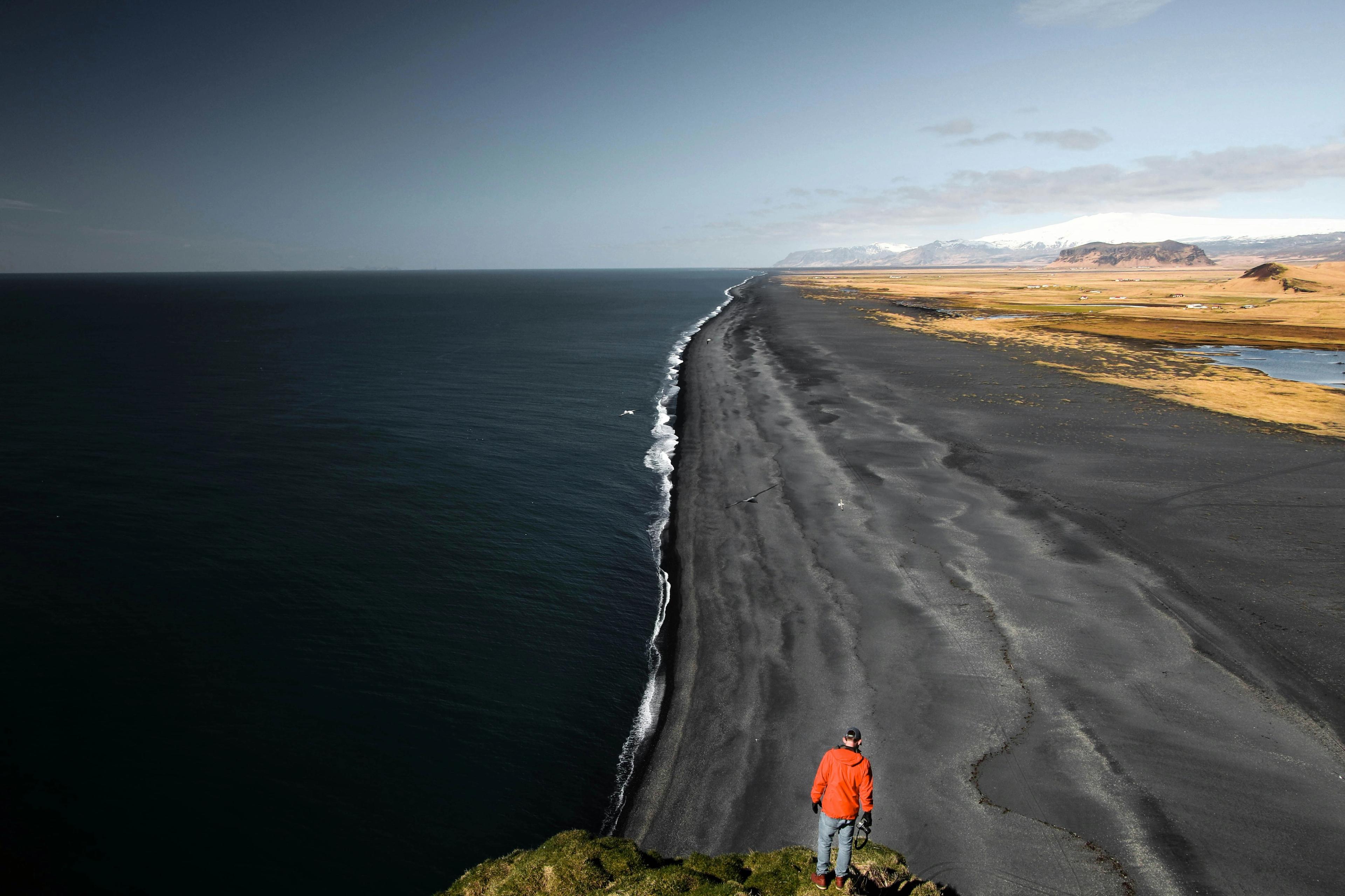 Person in an orange jacket on a grassy dune overlooks a black sand beach, ocean, and misty dark mountains.