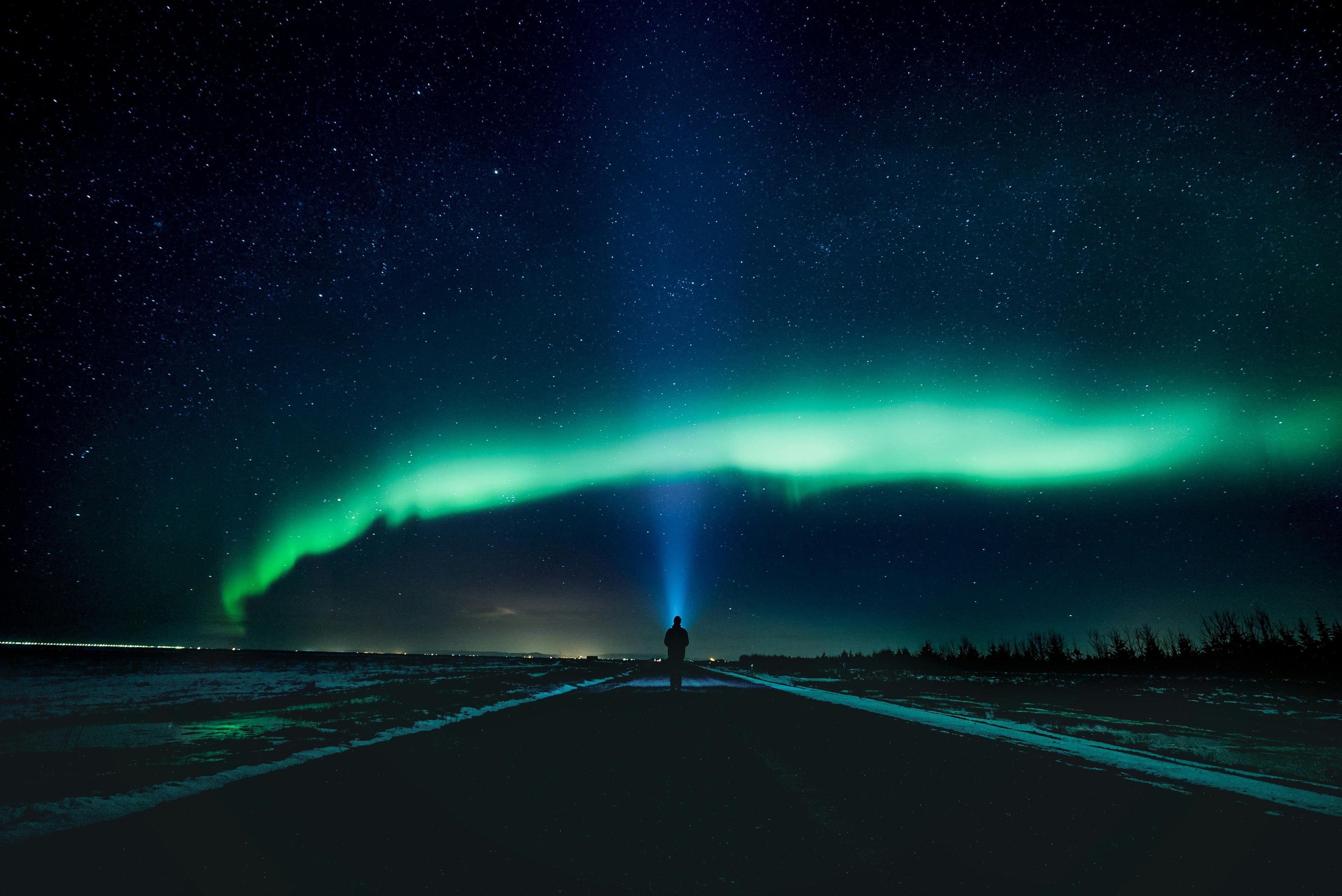 A lone figure stands on a road, gazing up at the vibrant green aurora borealis swirling in the night sky.