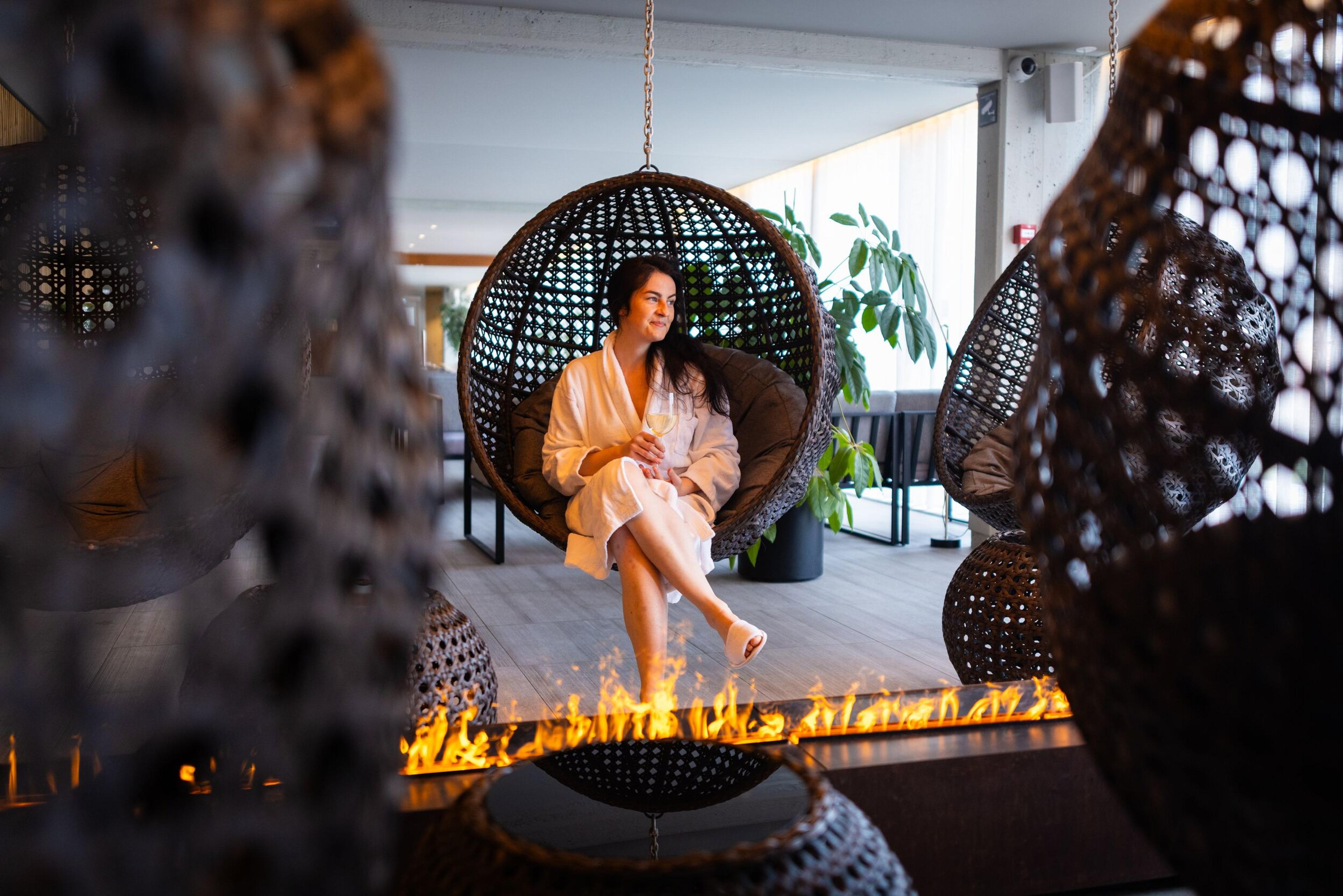 A woman relaxing in a hanging wicker chair at Krauma Bath, wearing a white robe and holding a glass of wine, with a modern fire feature and lush indoor greenery creating a serene spa atmosphere.