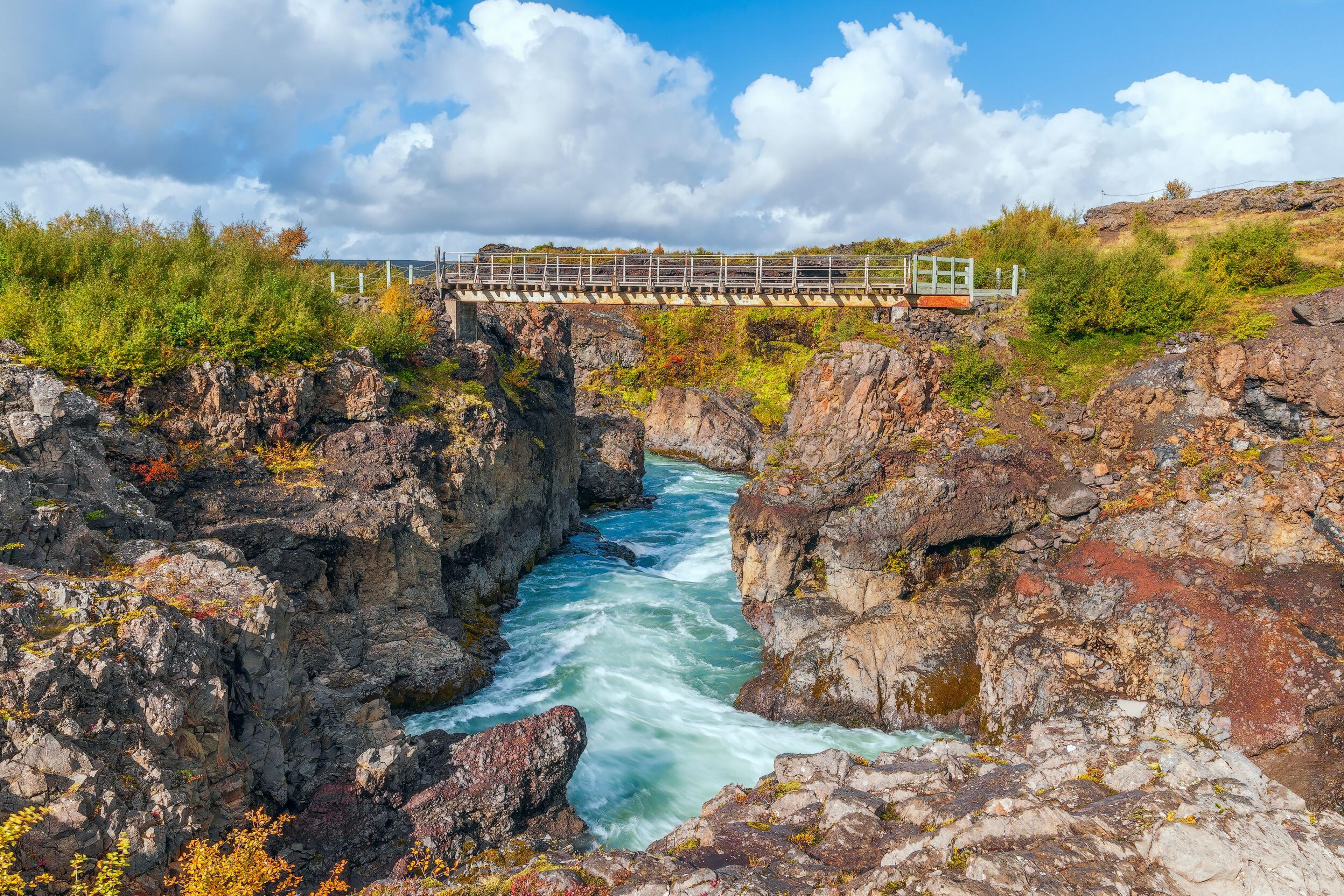 Barnafoss: a narrow canyon with a rushing blue river spanned by a wooden pedestrian bridge, framed by rocky cliffs and autumn foliage under a partly cloudy sky on the Silver Circle route Iceland.