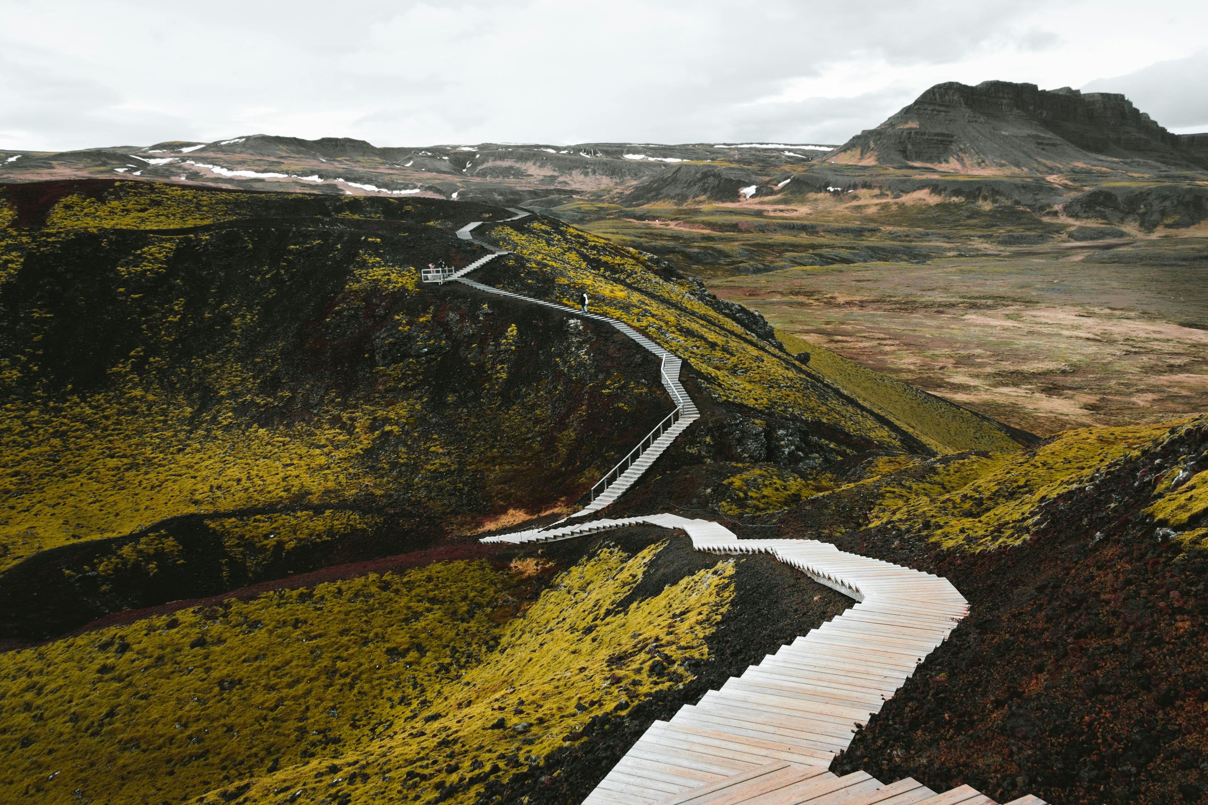 Wooden pathways winding along the vibrant mossy terrain of Gábók Crater, surrounded by rugged Icelandic landscapes under a cloudy sky.