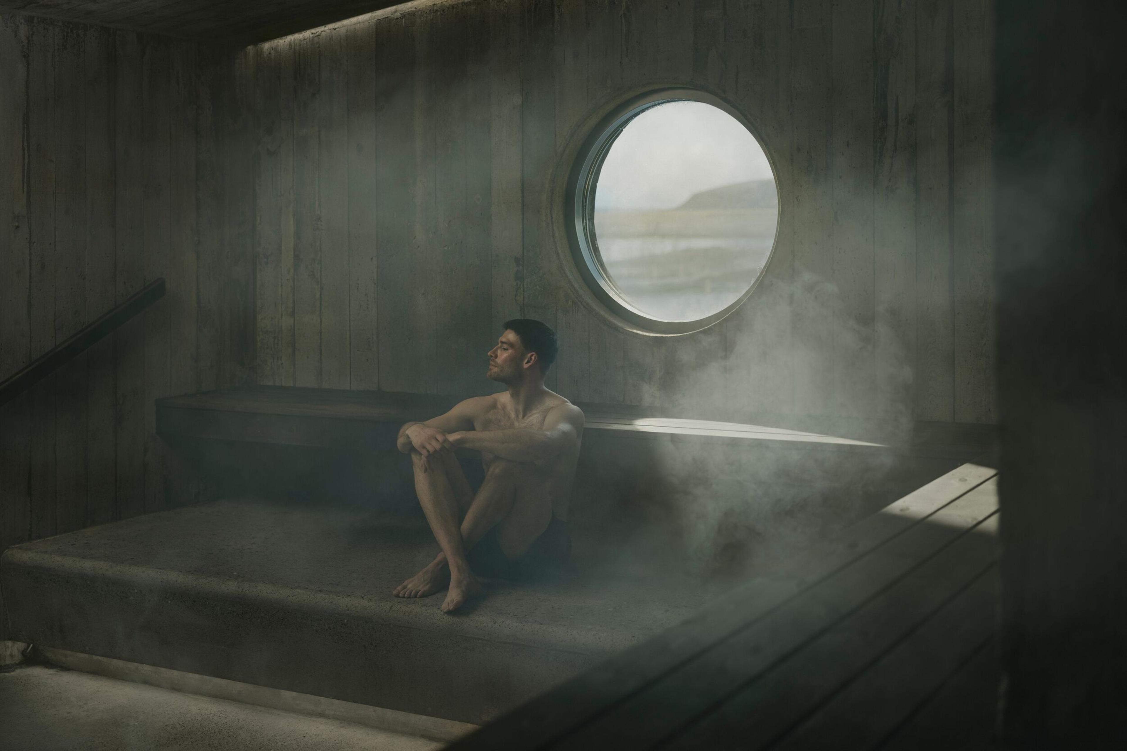 A man sits shirtless in a steamy room, looking out a round window at a natural landscape.