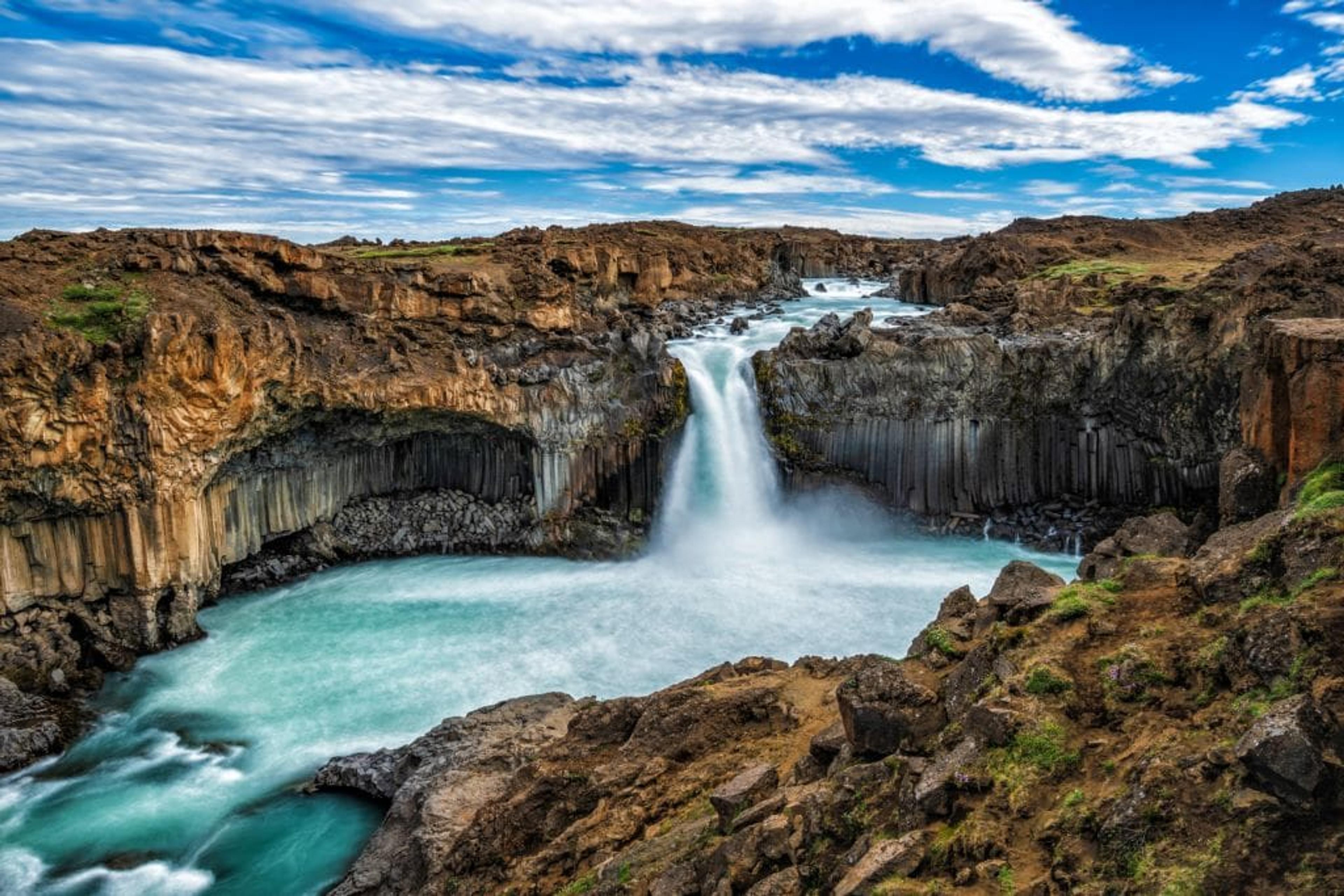 A waterfall cascades into a turquoise river surrounded by dark, rocky cliffs with prominent columnar basalt formations under a partly cloudy sky.