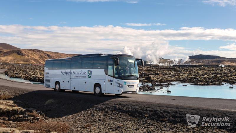 A white 'Reykjavik Excursions' bus on a road in a volcanic landscape with blue water and a geothermal plant emitting steam.