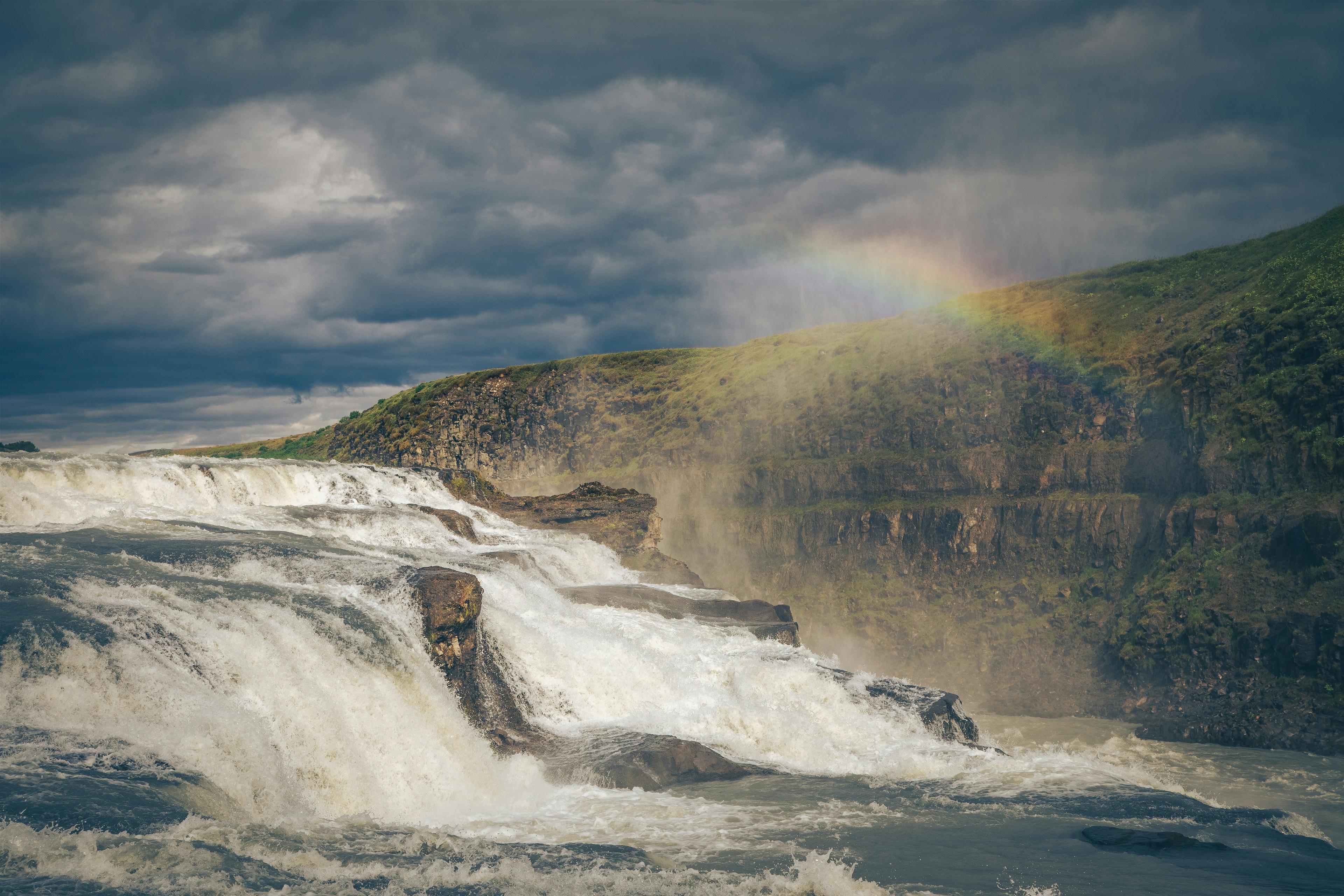 Gullfoss waterfall cascading into a river within a rugged landscape, bathed in sunlight piercing through the mist, a scene characteristic of the Golden Circle tourist route in Iceland.