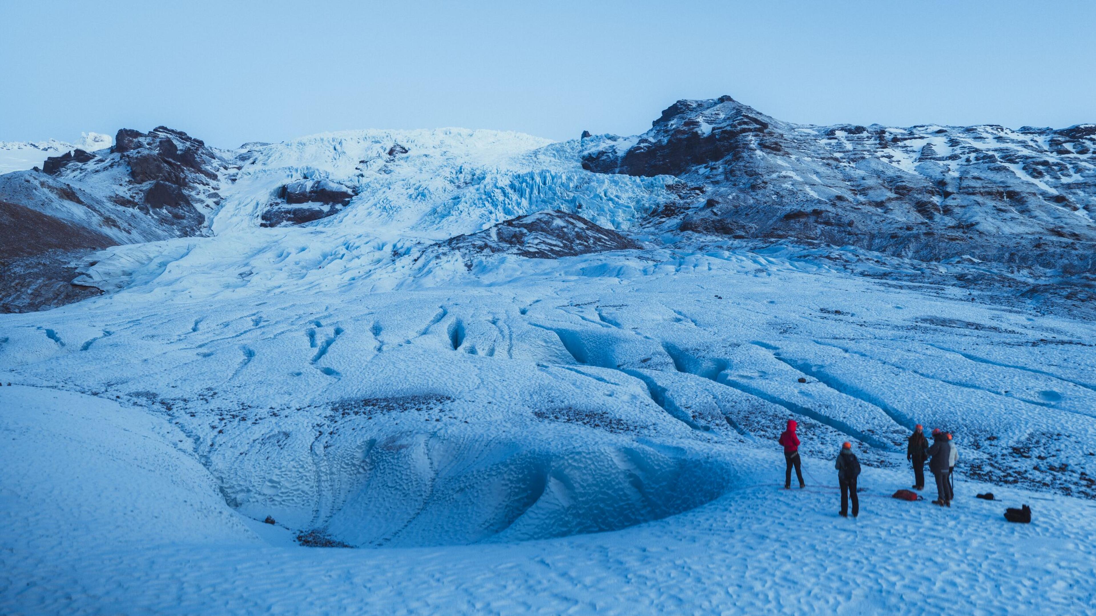 Four people on a large glacier with snowy mountains in the background.