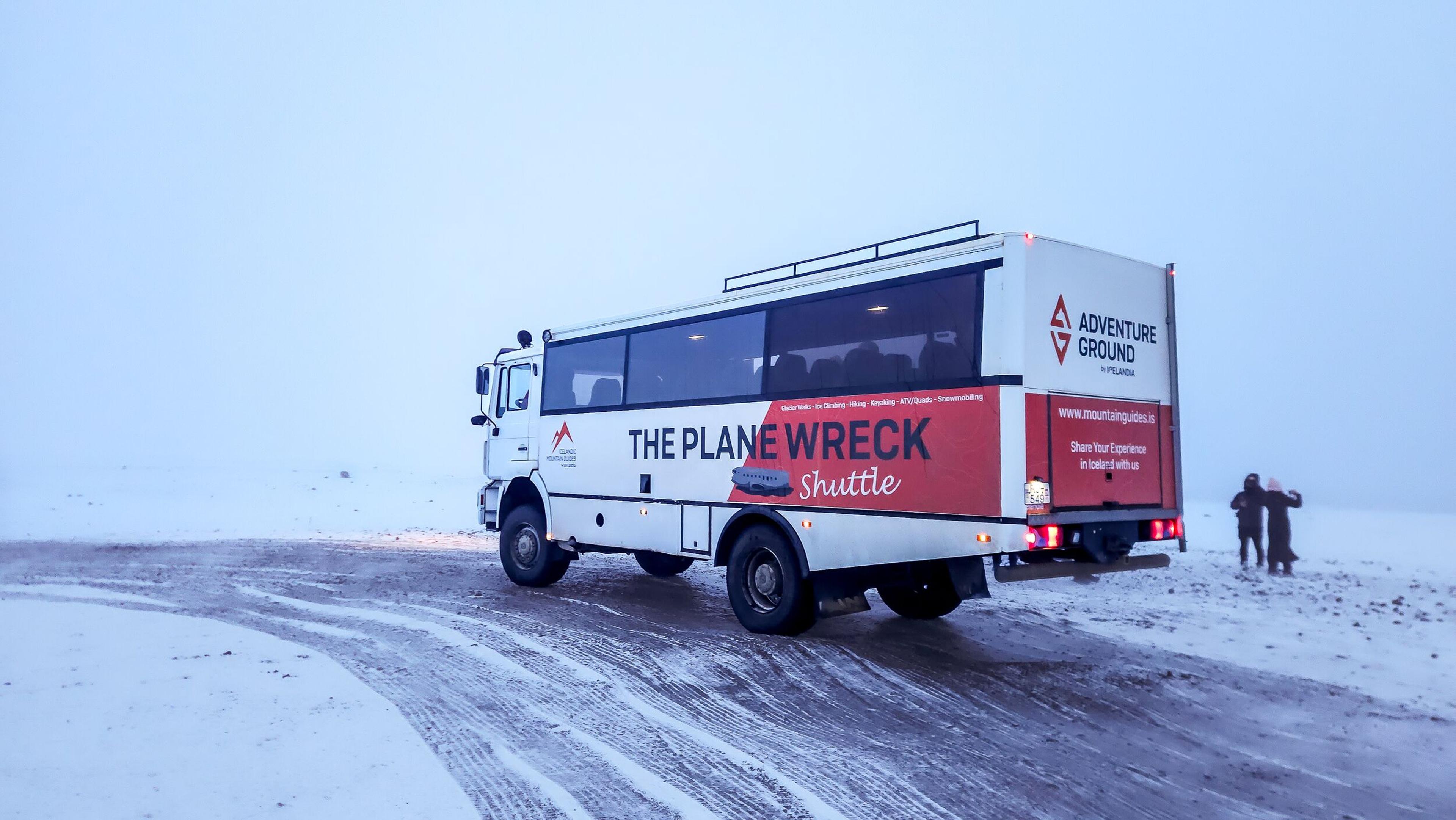 A white "The Plane Wreck Shuttle" bus on a snowy, desolate landscape with two figures in the distance.