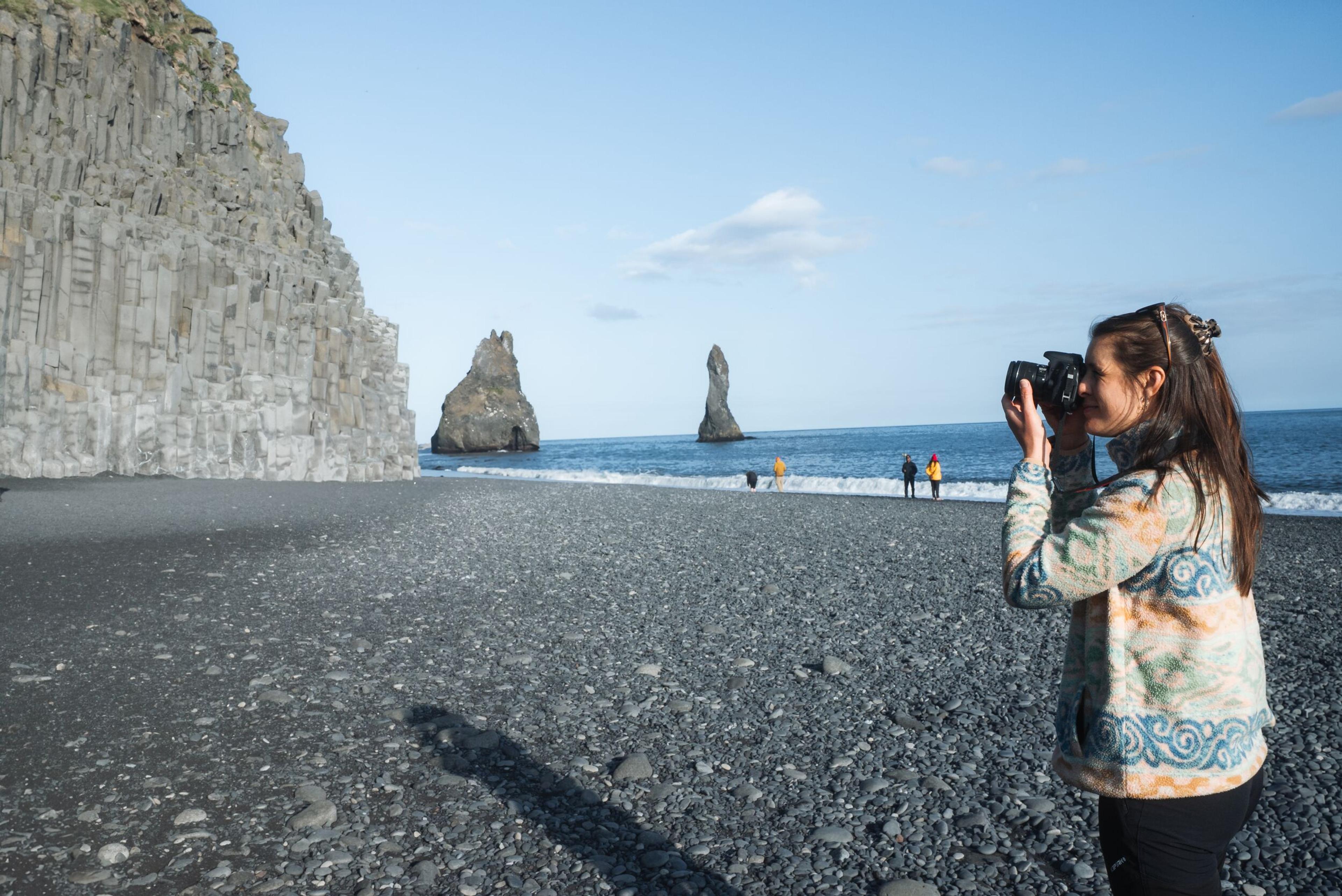 Woman photographing a black pebble beach with basalt columns and sea stacks.