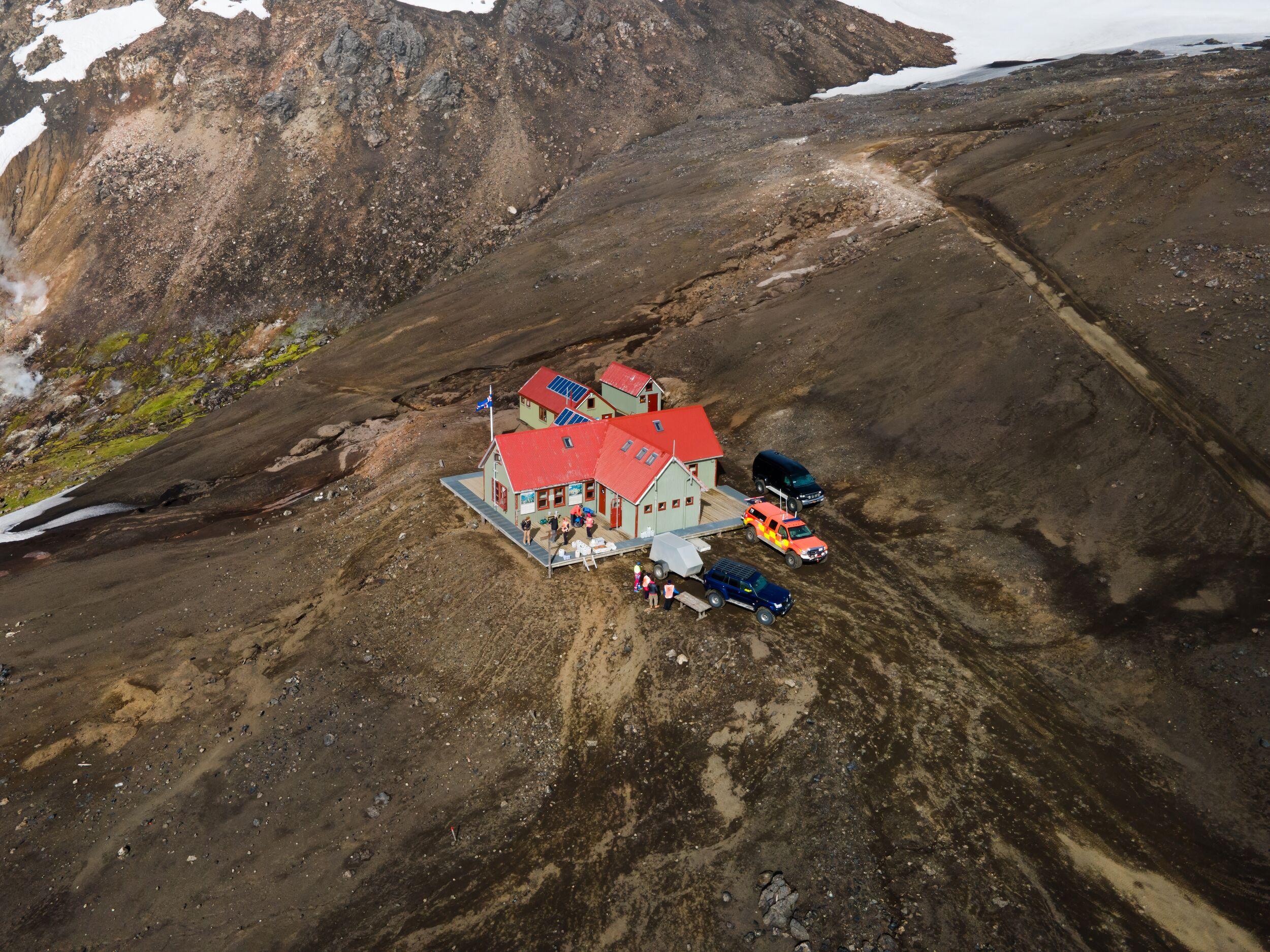 Otherworldly Landscape: Hrafntinnusker Hut Amidst Brown Terrain with Steam Rising, Snow-Dusted Hills, and Parked Trucks