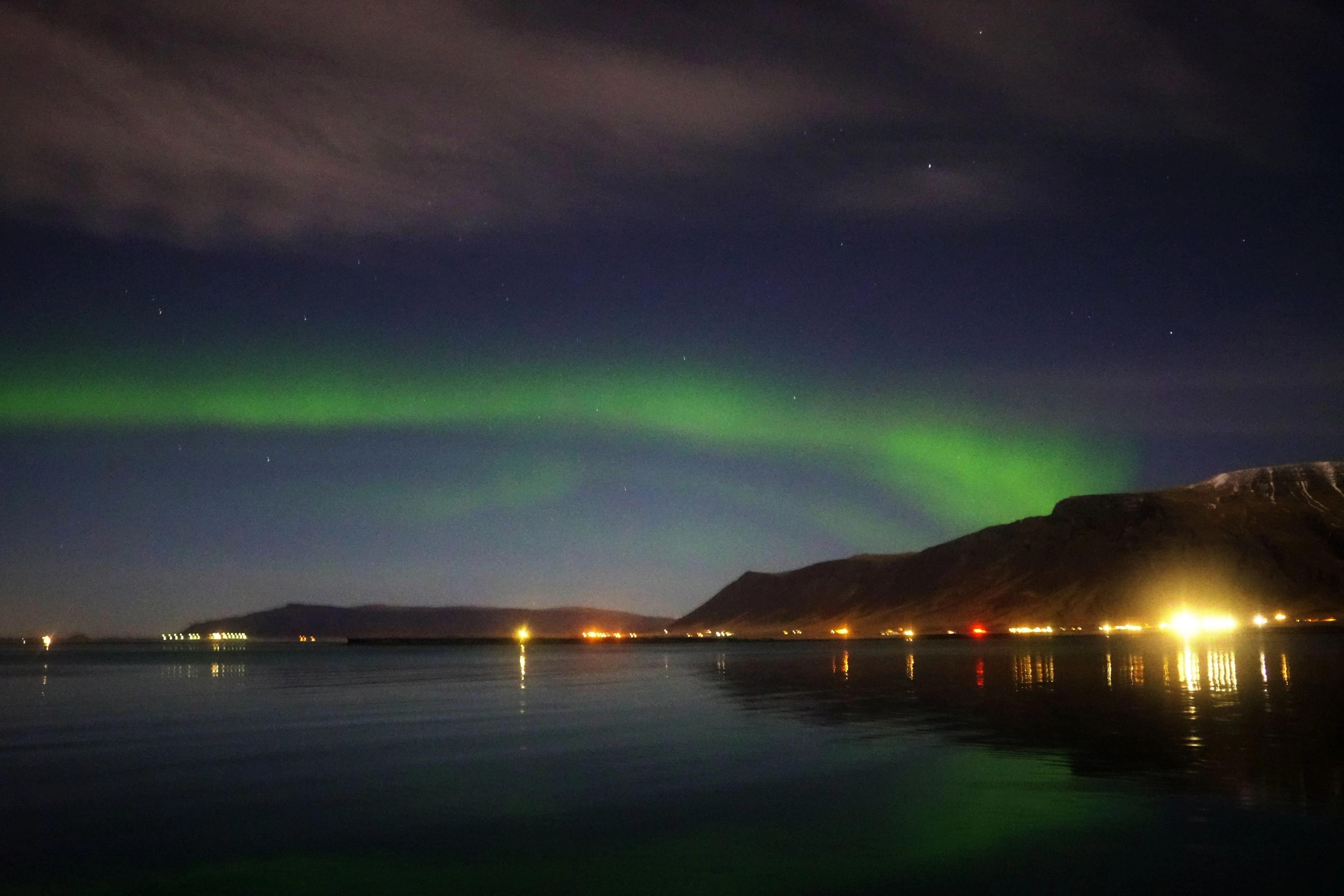 Green aurora borealis over dark water reflecting town lights and mountains.