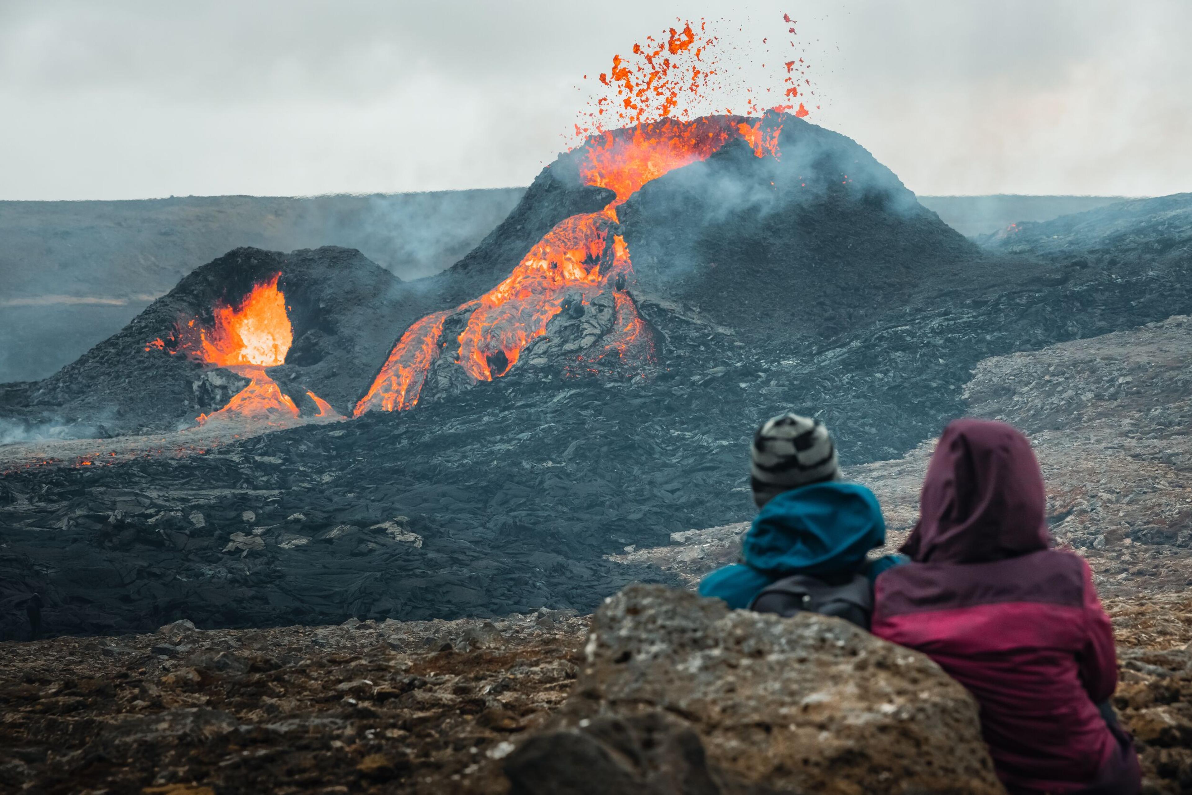 Visitors trekking across the rugged terrain of an Icelandic lava field, with an active volcano in the background erupting, spewing lava and ash into the air, a dynamic scene of nature's power.