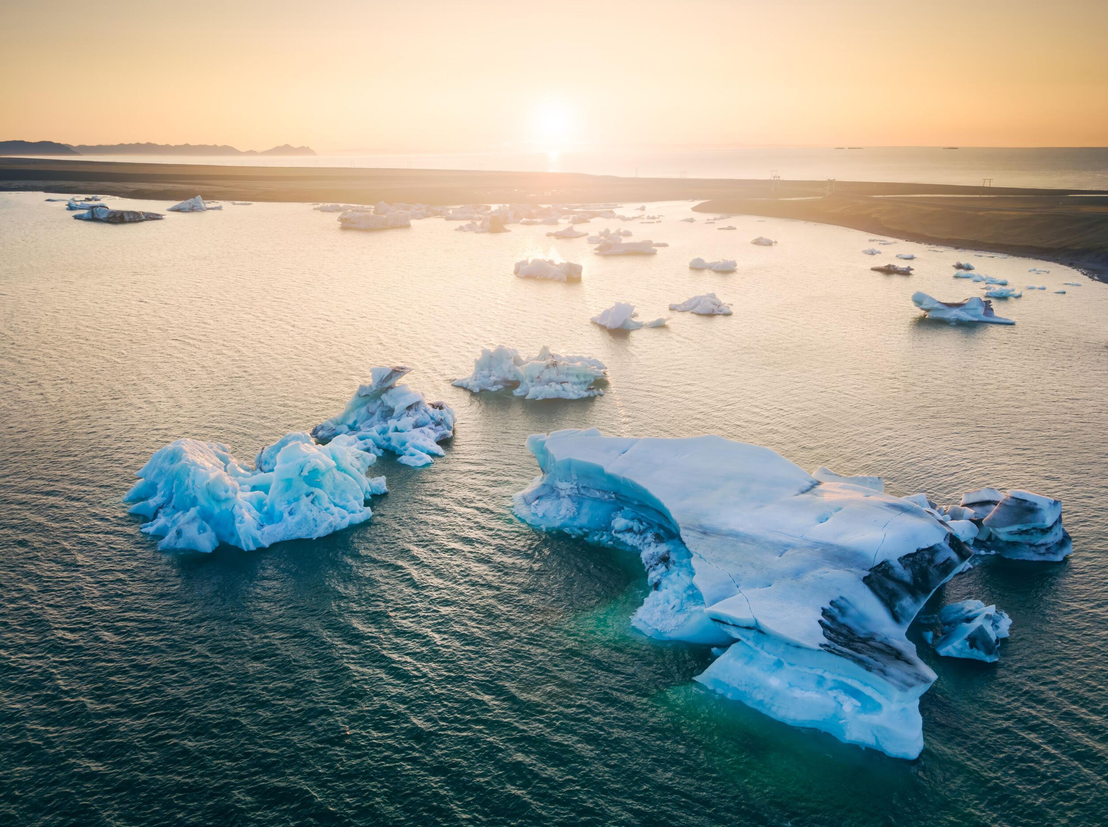 Aerial view of blue-tinged icebergs floating in a glacial lagoon at sunset, with the sun casting a warm glow over the tranquil water and a hint of distant land in the backdrop.