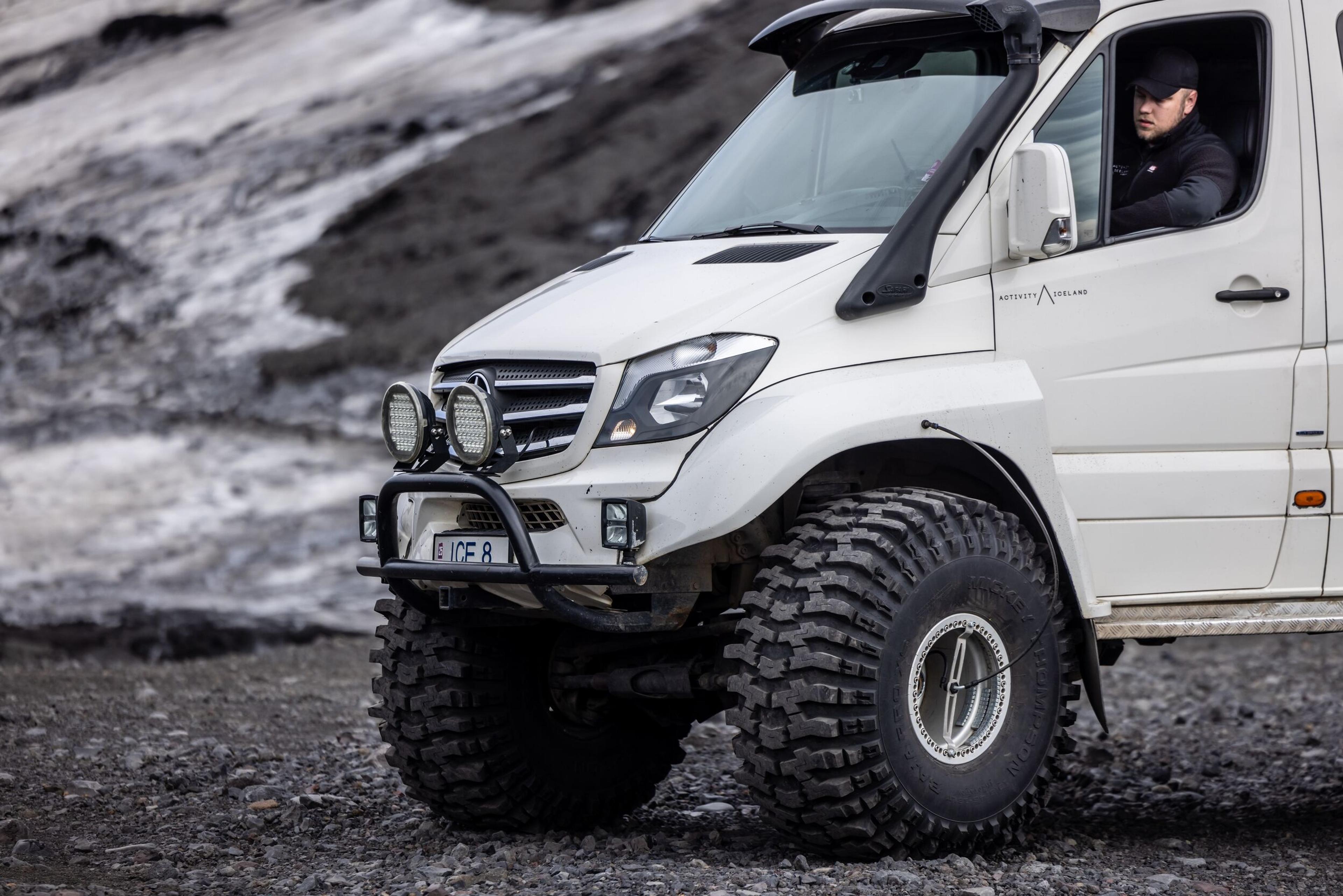  White modified 4x4 vehicle with large off-road tires, driving on rocky Icelandic terrain near a glacier backdrop.