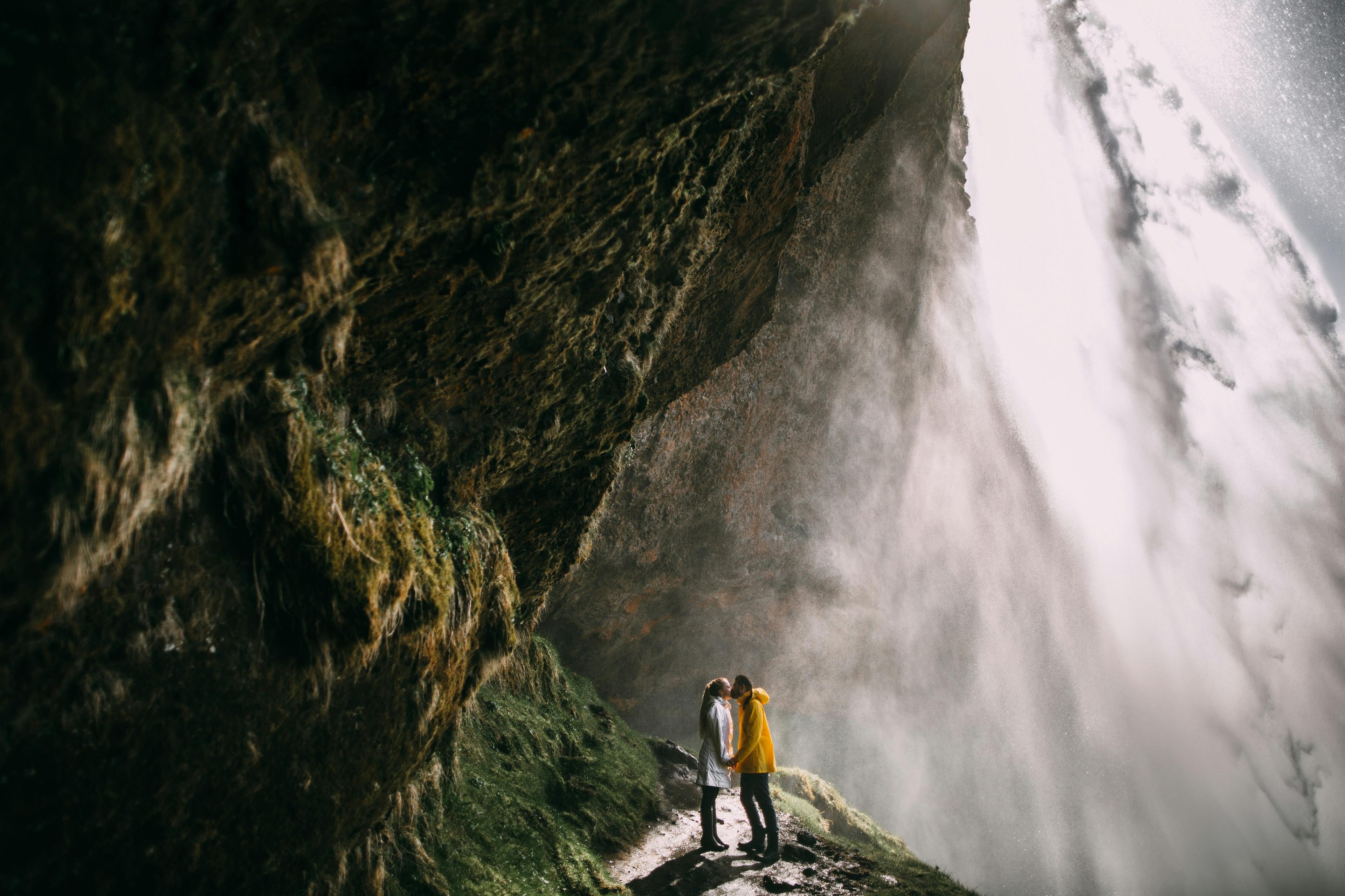 A couple stands close together behind the misty cascade of Seljalandsfoss waterfall, surrounded by lush, moss-covered cliffs.