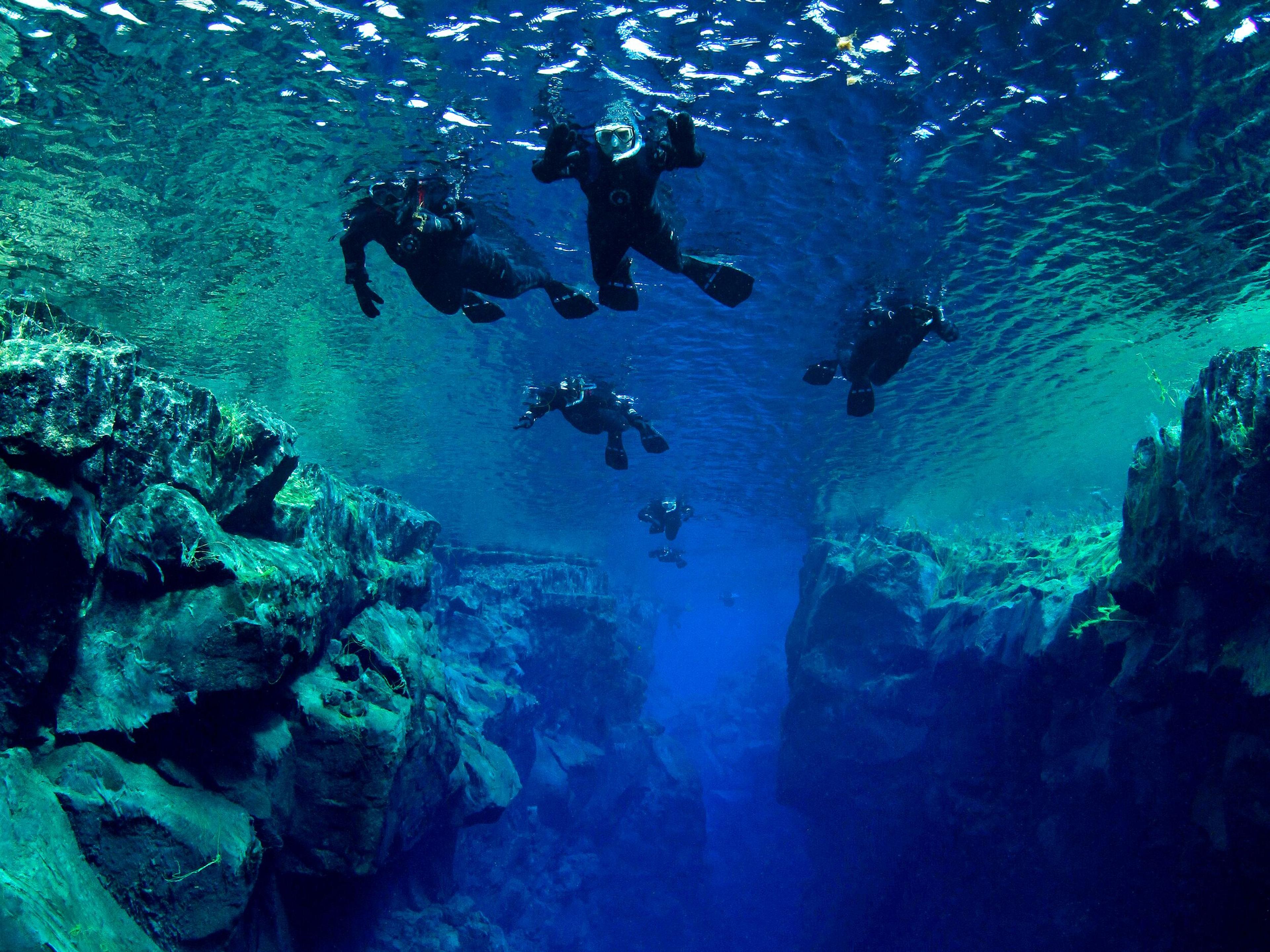 Several people snorkel through a crystal-clear underwater rift with rocky walls.