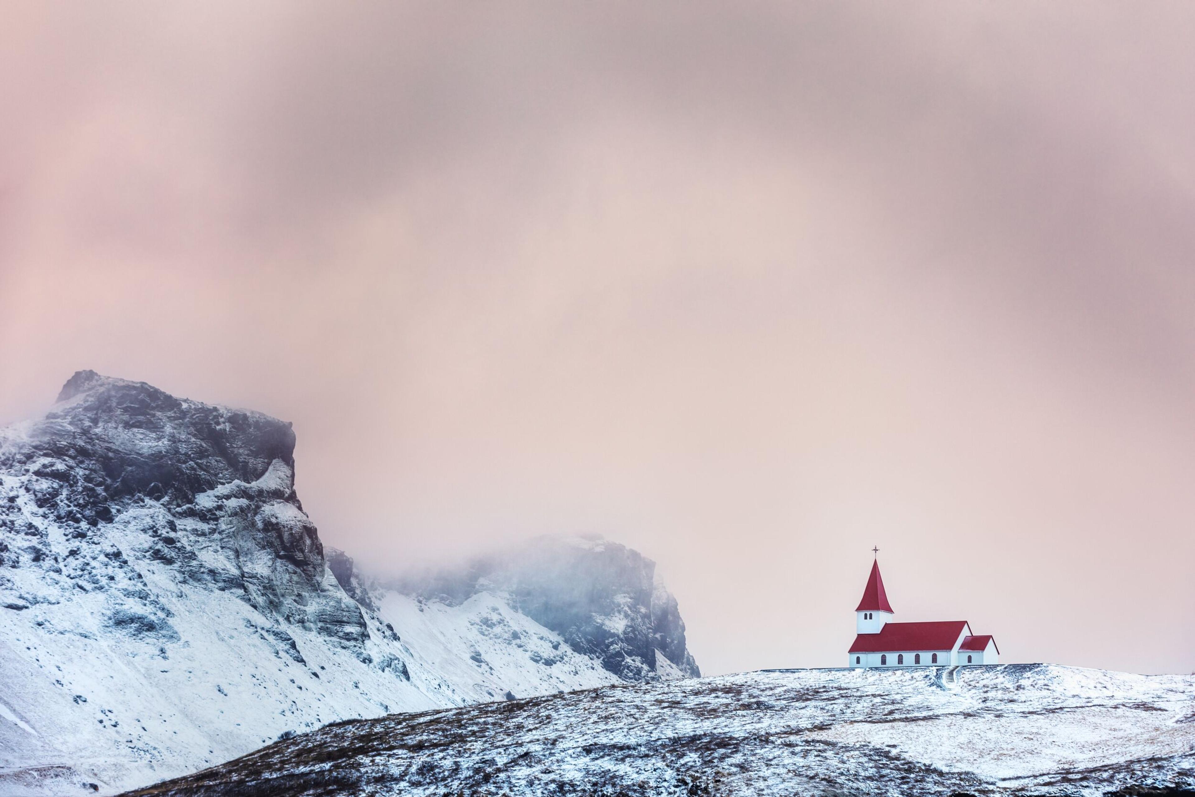 A small church with a bright red roof stands out against a snowy landscape, with rugged cliffs partially shrouded by mist in the background. The sky is a soft pink hue, adding a serene atmosphere to the scene.