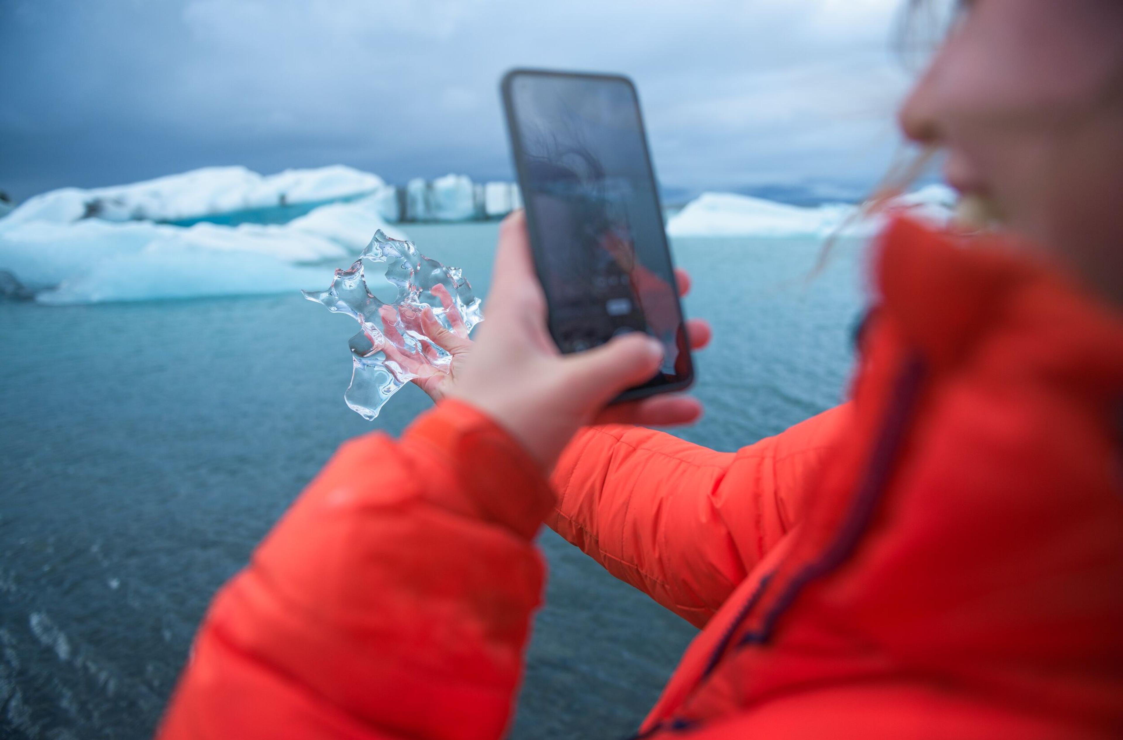Individual at a glacier lagoon, holding a chunk of ice and capturing it with her phone