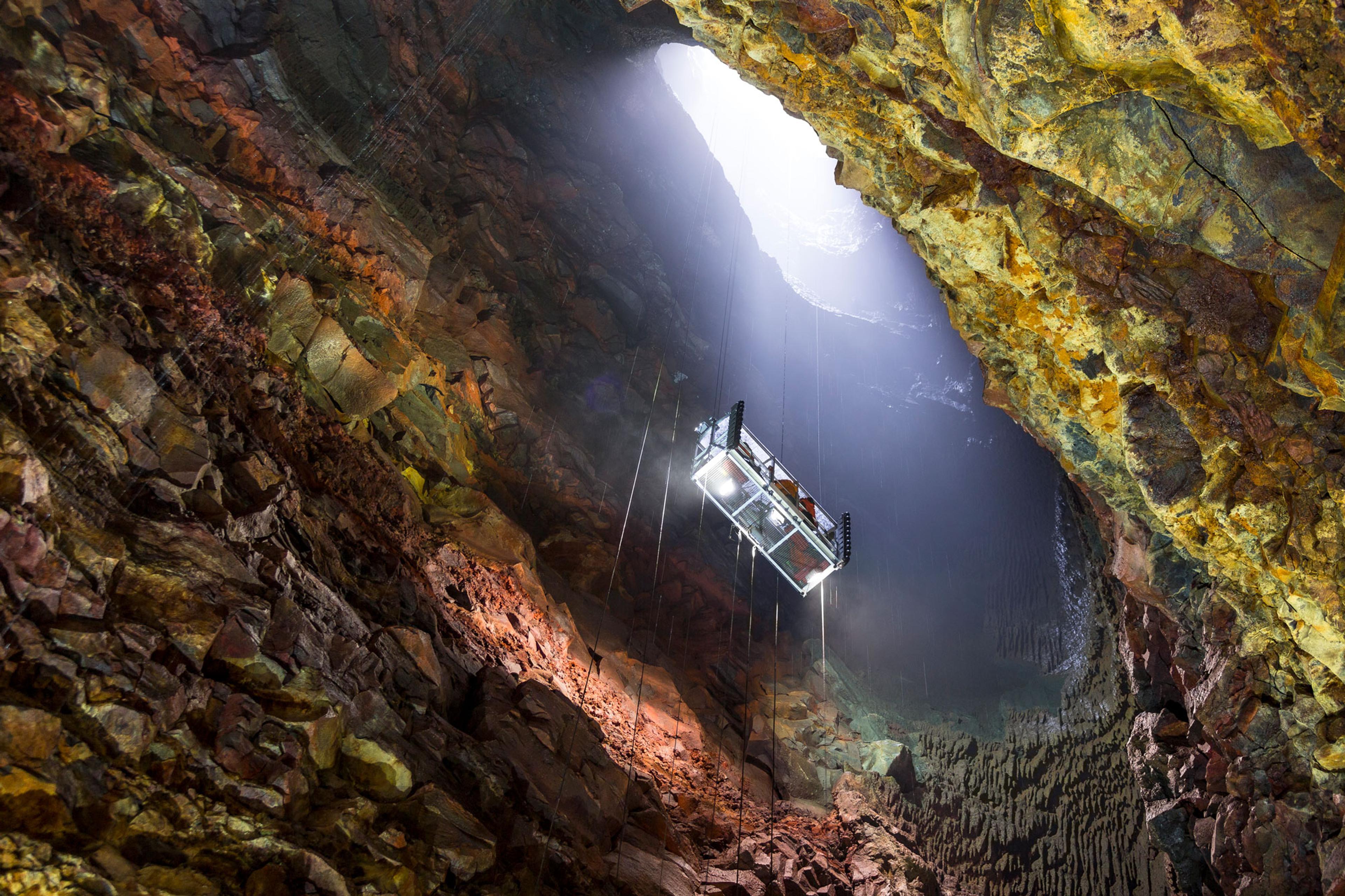 A metal elevator cage descends into a large, colorful volcanic cavern, illuminated by light from above.
